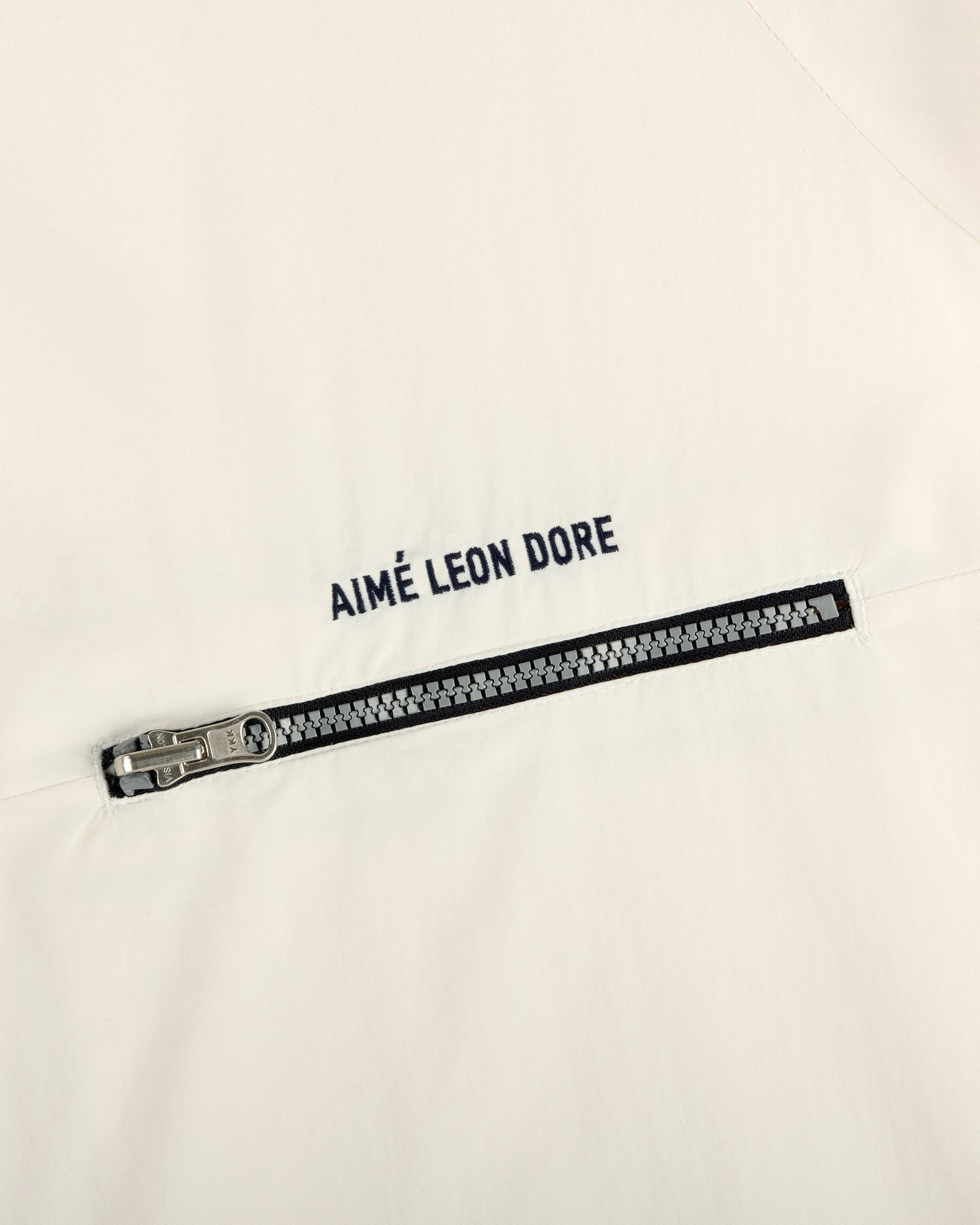 Close-up of an off-white Nylon Packable Windbreaker with a black zipper and AIMÉ LEON DORE text above the zipper.