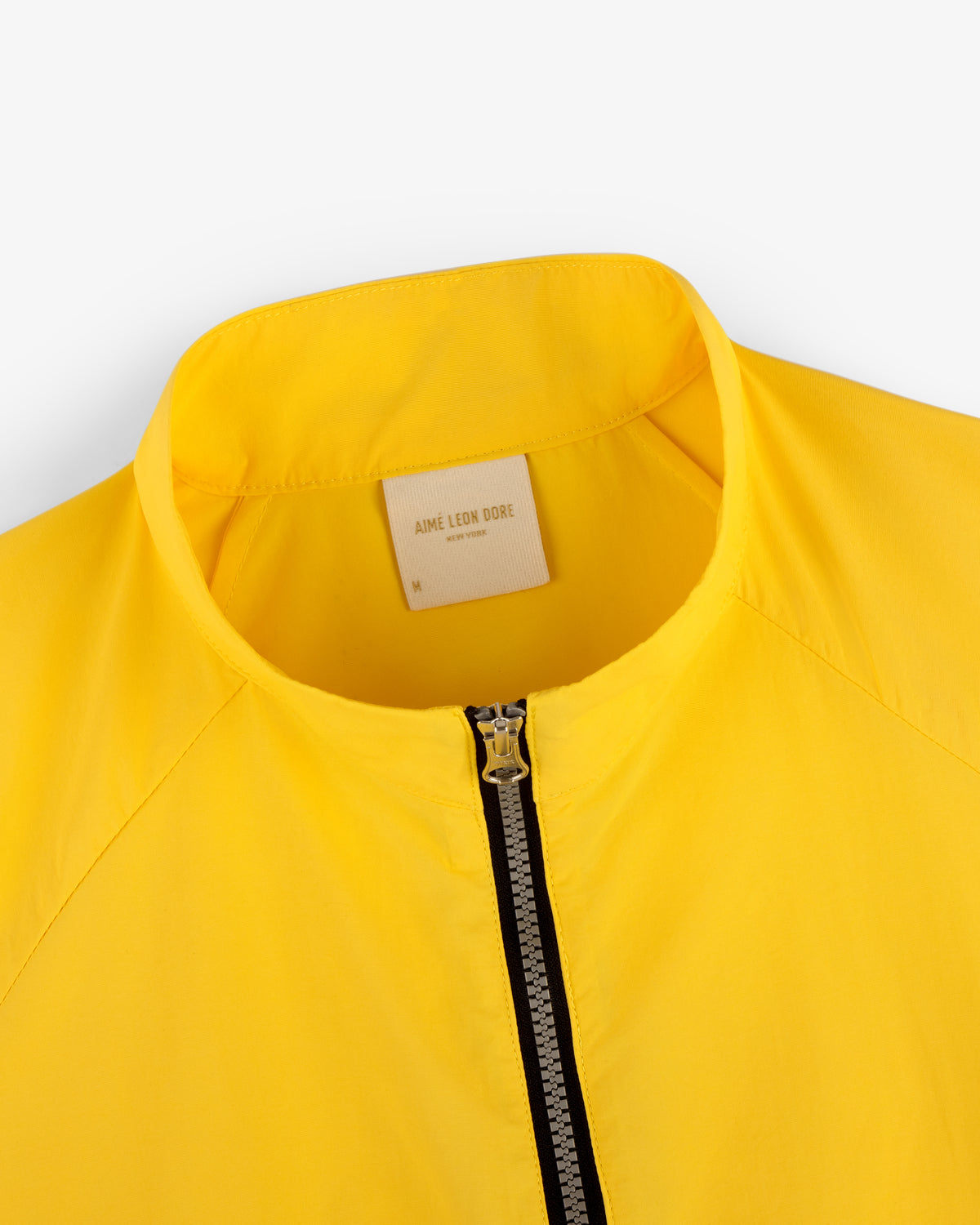 Close-up of a bright yellow Nylon Packable Windbreaker with a zipper and an Aimé Leon Dore label on the inside collar.