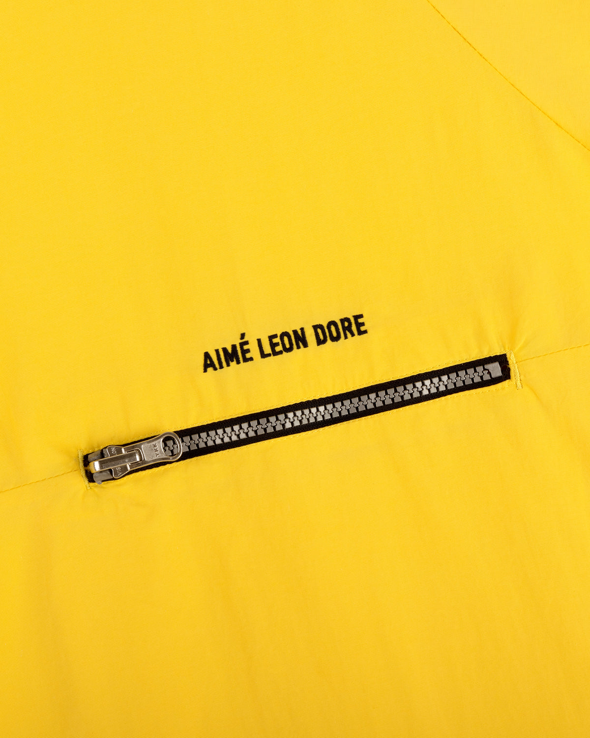 Close-up of a yellow Nylon Packable Windbreaker fabric with a diagonal zipper and AIMÉ LEON DORE text above it.