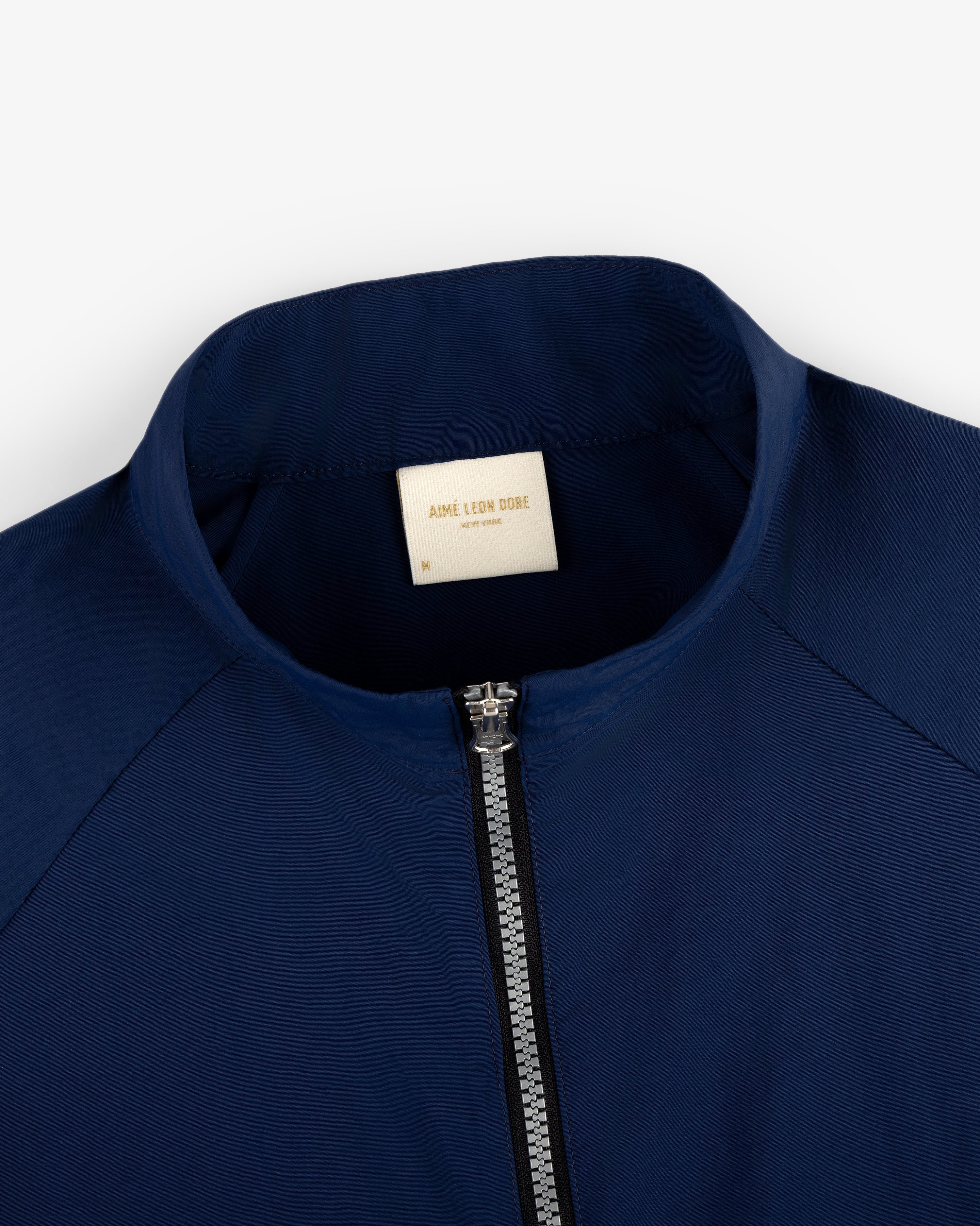 Close-up of the Nylon Packable Windbreaker in navy blue, featuring a silver zipper and AIMÉ LEON DORE collar label.