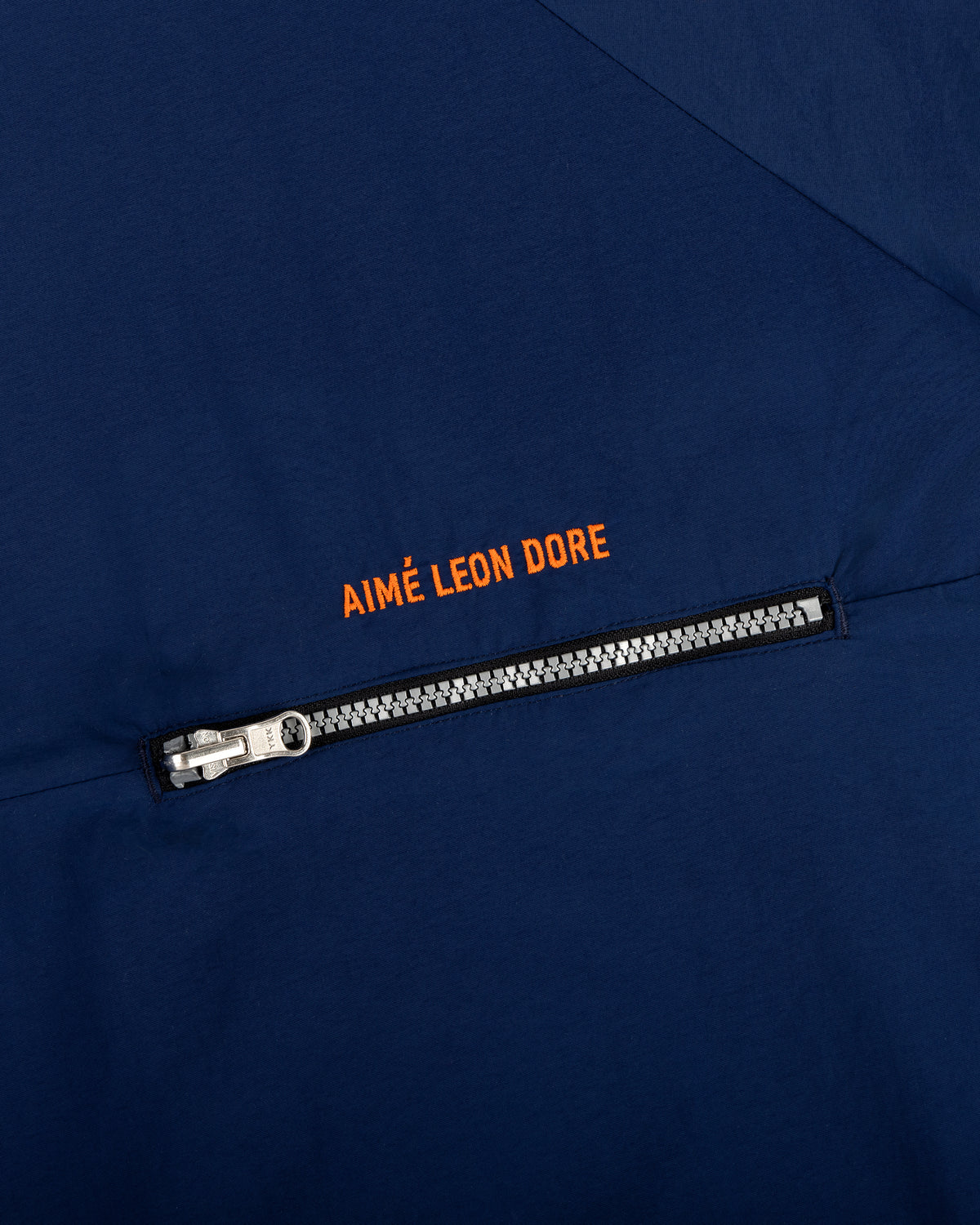 Close-up of a navy Nylon Packable Windbreaker with a silver zipper and Aimé Leon Dore in orange above the zipper.