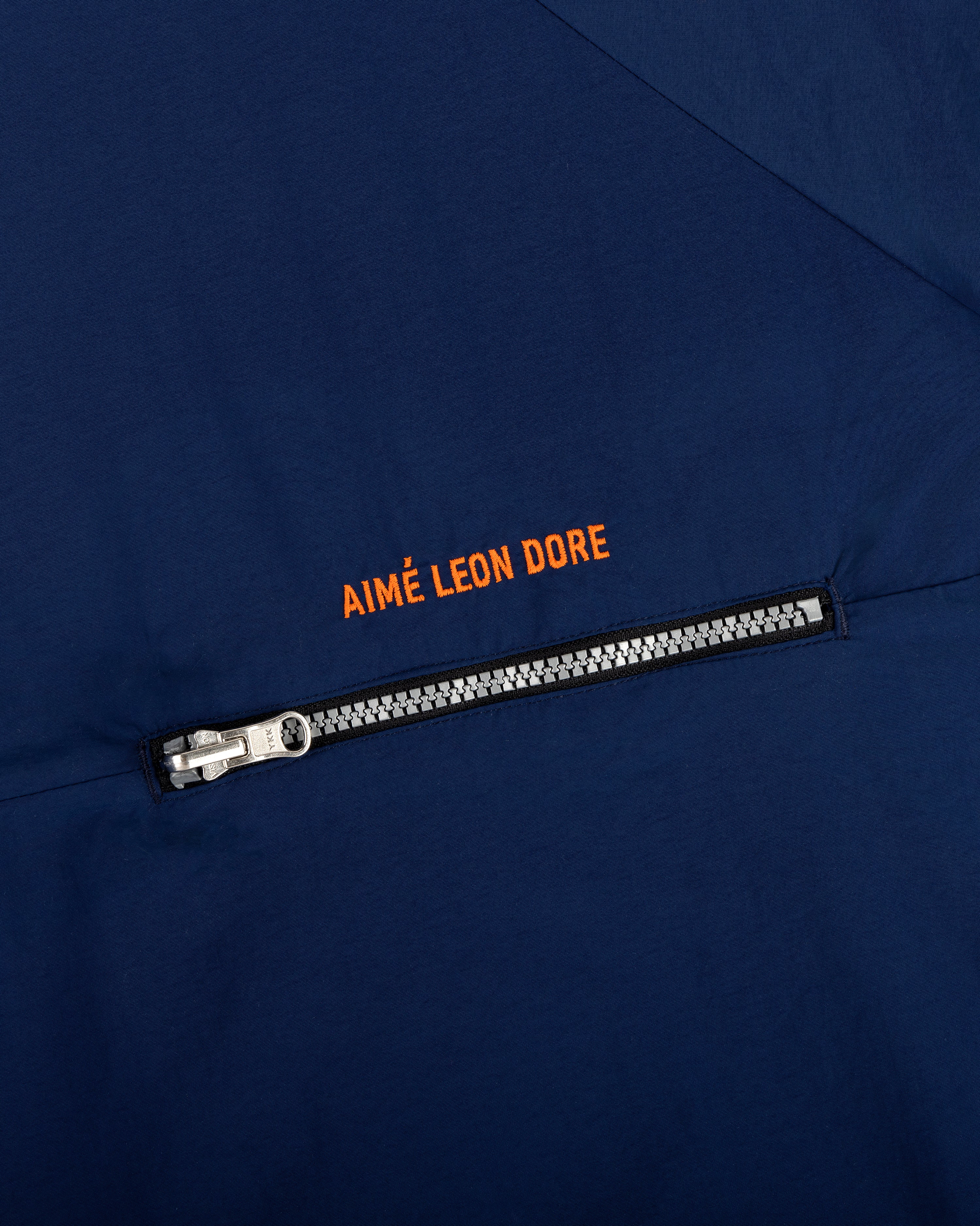 Close-up of a navy Nylon Packable Windbreaker with a silver zipper and Aimé Leon Dore in orange above the zipper.