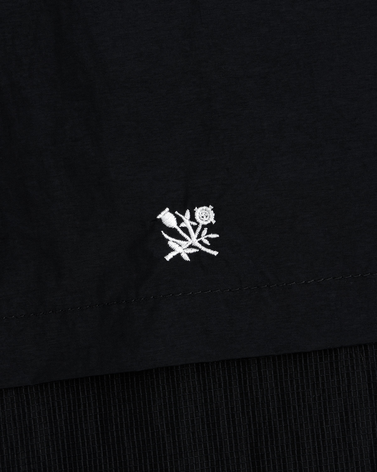 Small white embroidered rose with leaves on black Nylon Track Jacket, near the hem.