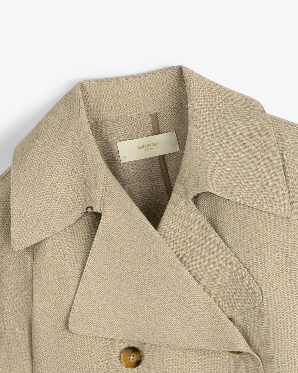 Close-up of the Linen Trench Coat in beige with wide lapels, button, and inner collar label visible.