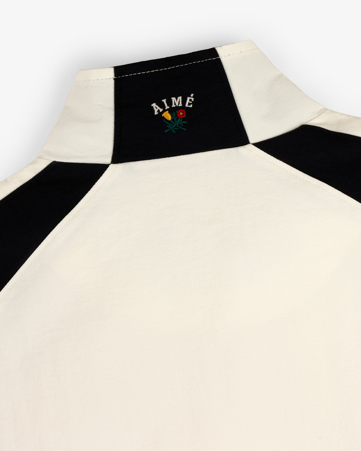 Close-up of a cream and black Crest Nylon Half-Zip Pullover with AIMÉ and a small embroidered flower on the back neck.