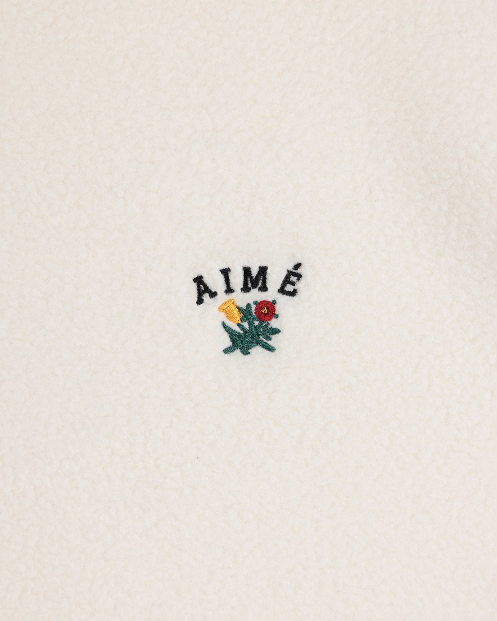 Crest Quarter-Zip Fleece Pullover with AIMÉ text and a small yellow and red flower embroidered on white fleece.