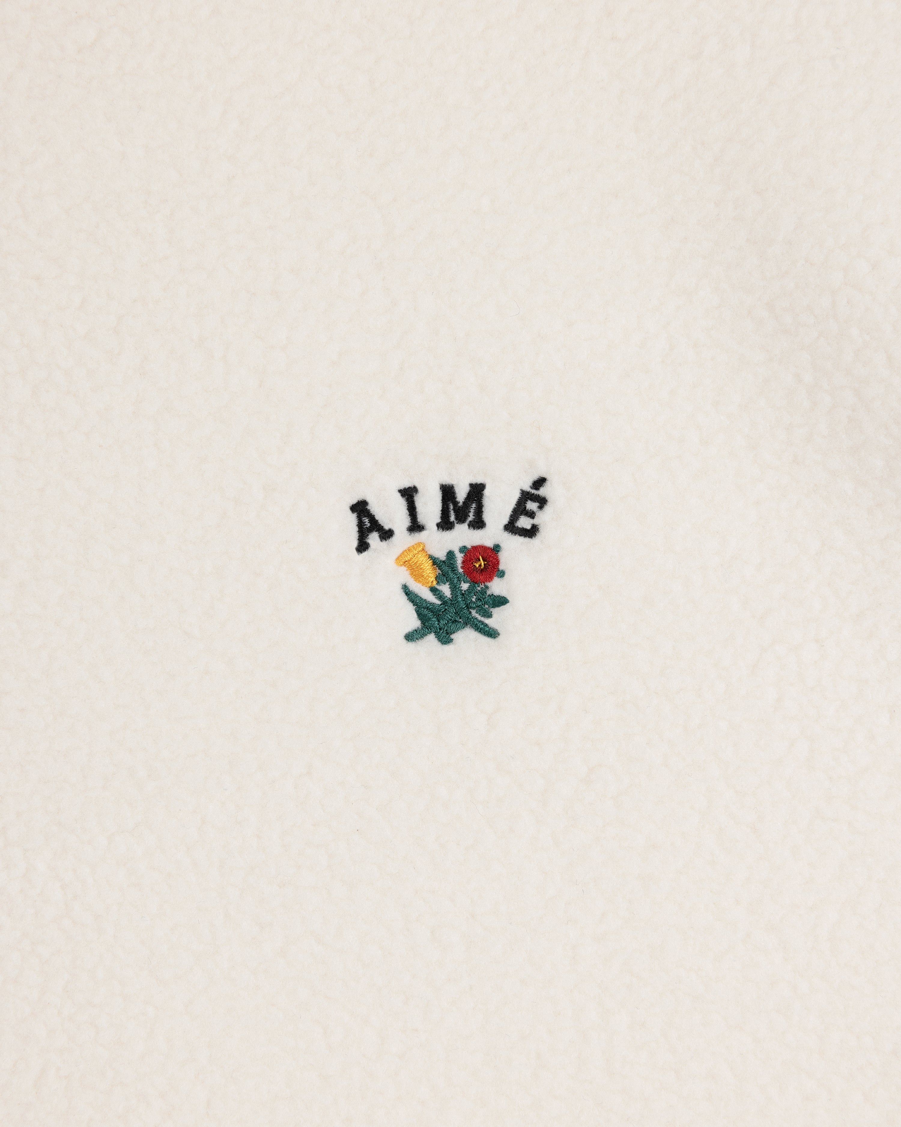 Crest Quarter-Zip Fleece Pullover with AIMÉ text and a small yellow and red flower embroidered on white fleece.