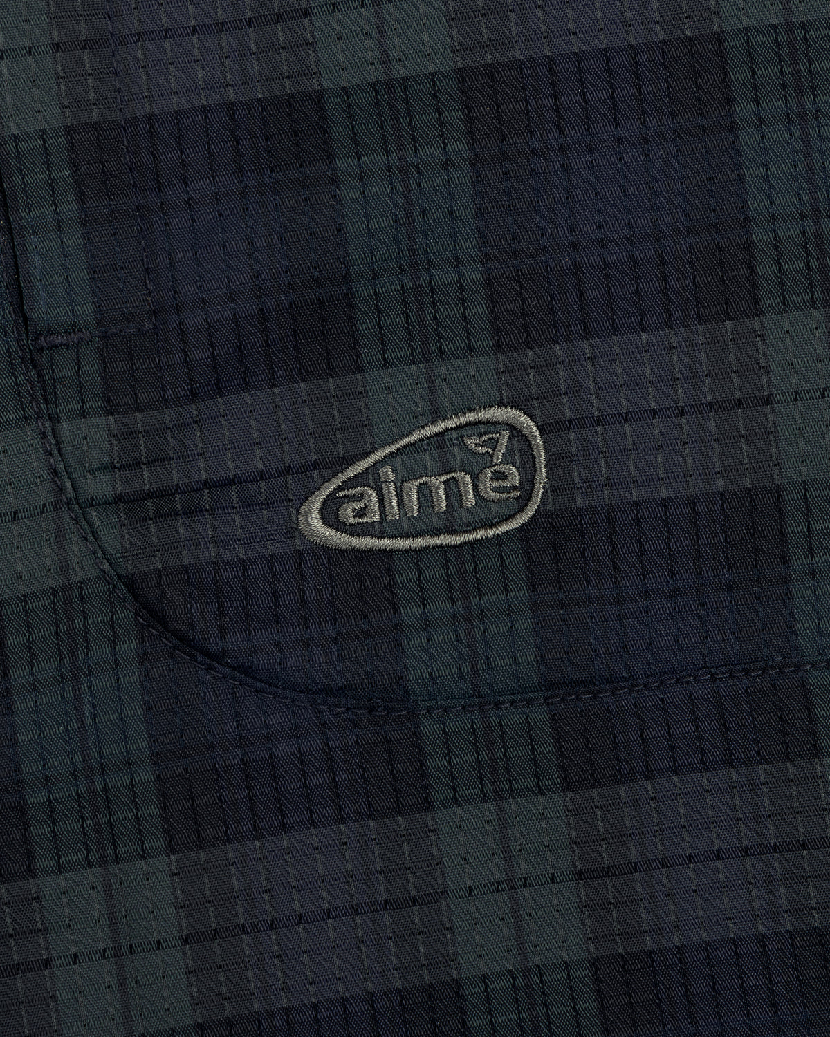 Close-up of ALD Golf Lightweight Tips Windbreaker showing dark plaid fabric, stitched pocket, and light aime logo.