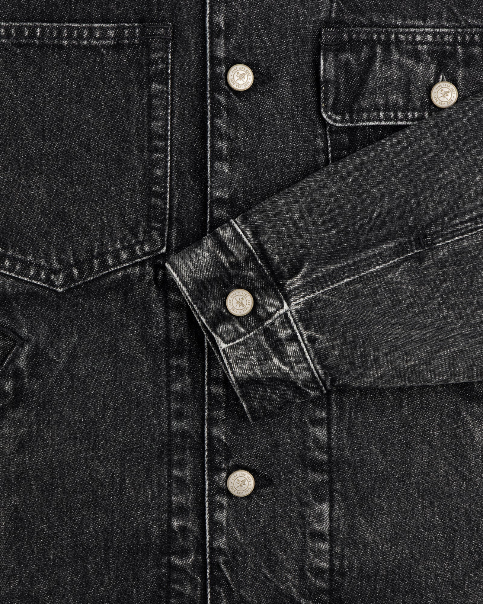 Close-up of a Denim Trucker Jacket in black with silver buttons, chest pockets, and one sleeve across the front.