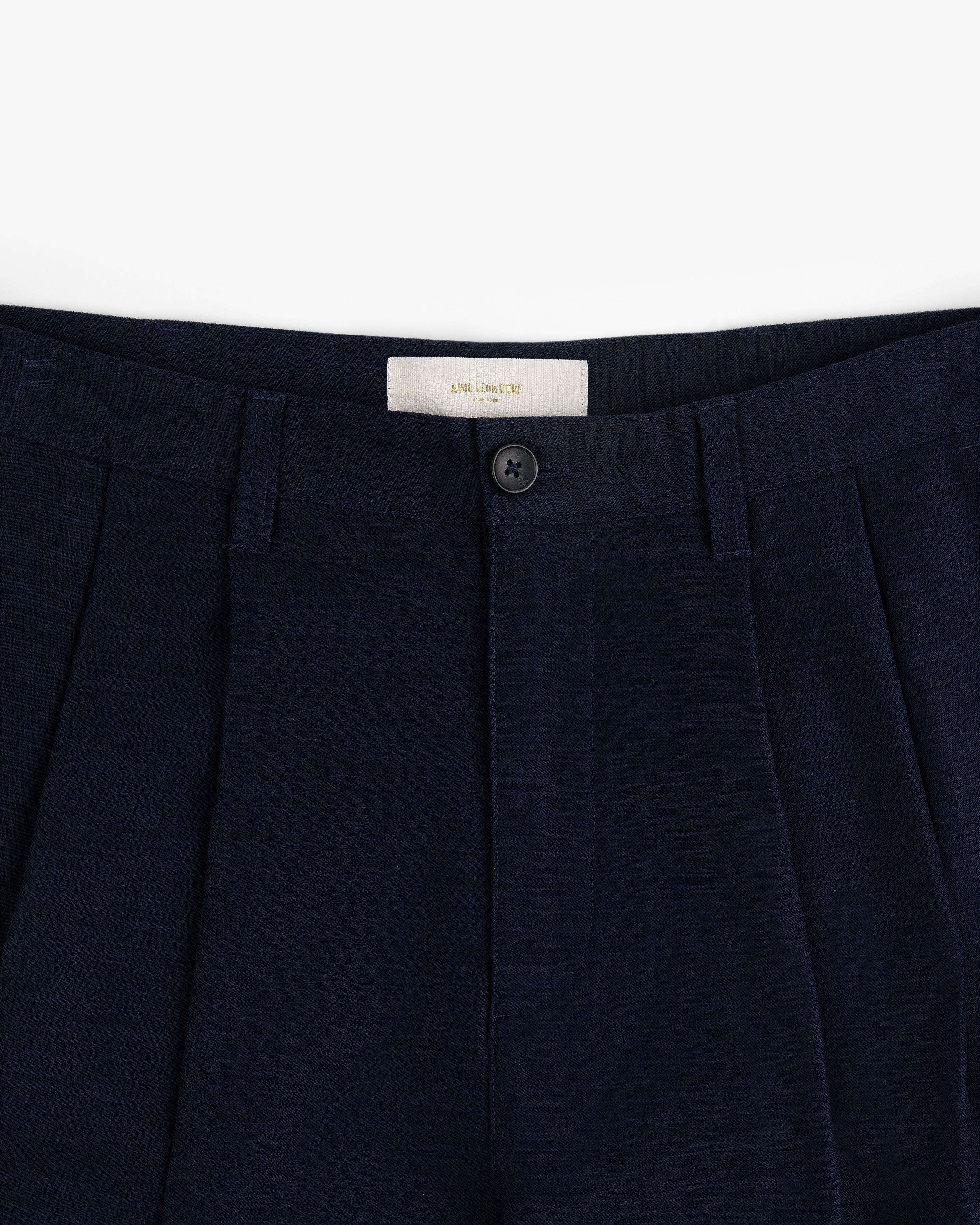 Double Pleated Pant: Navy blue dress pants with waistband, belt loops, pleats, and a black button above the zipper.