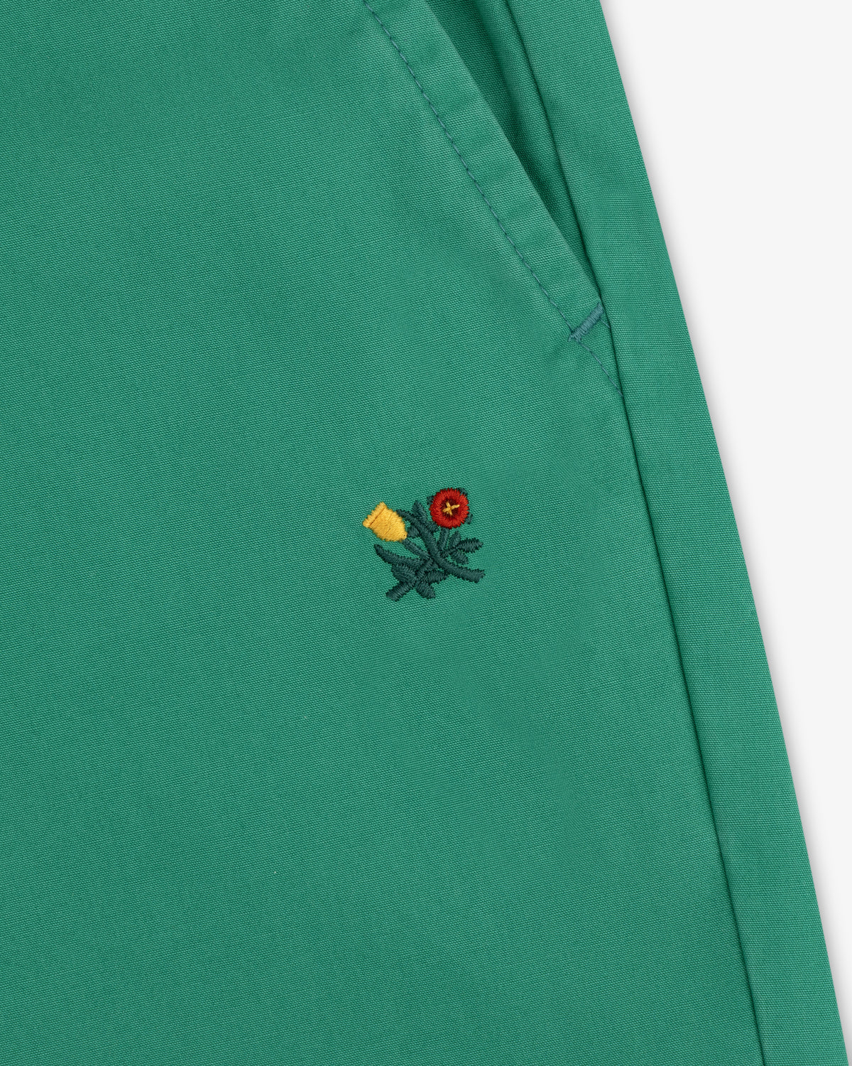 Washed Cotton Windbreaker Pant in green with pocket and small embroidered flower—red center, yellow petals, green stem.