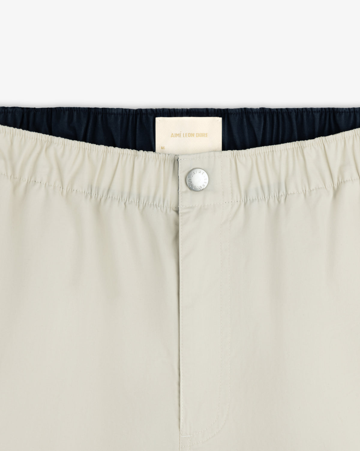 Close-up of Washed Cotton Windbreaker Pant in beige with elastic waistband, button closure, and Aimé Leon Dore label.
