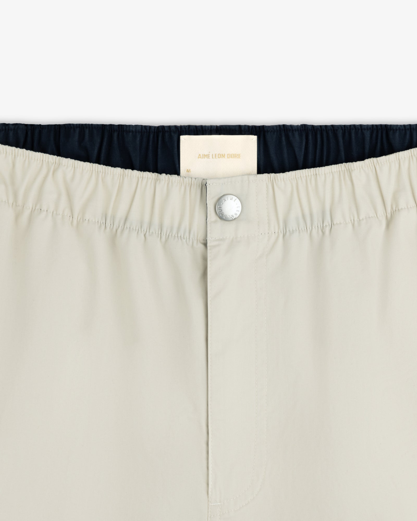Close-up of Washed Cotton Windbreaker Pant in beige with elastic waistband, button closure, and Aimé Leon Dore label.