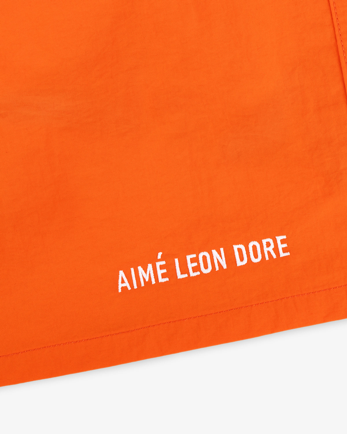 Close-up of orange Logo Swim Short with AIMÉ LEON DORE embroidered in white near the hem.