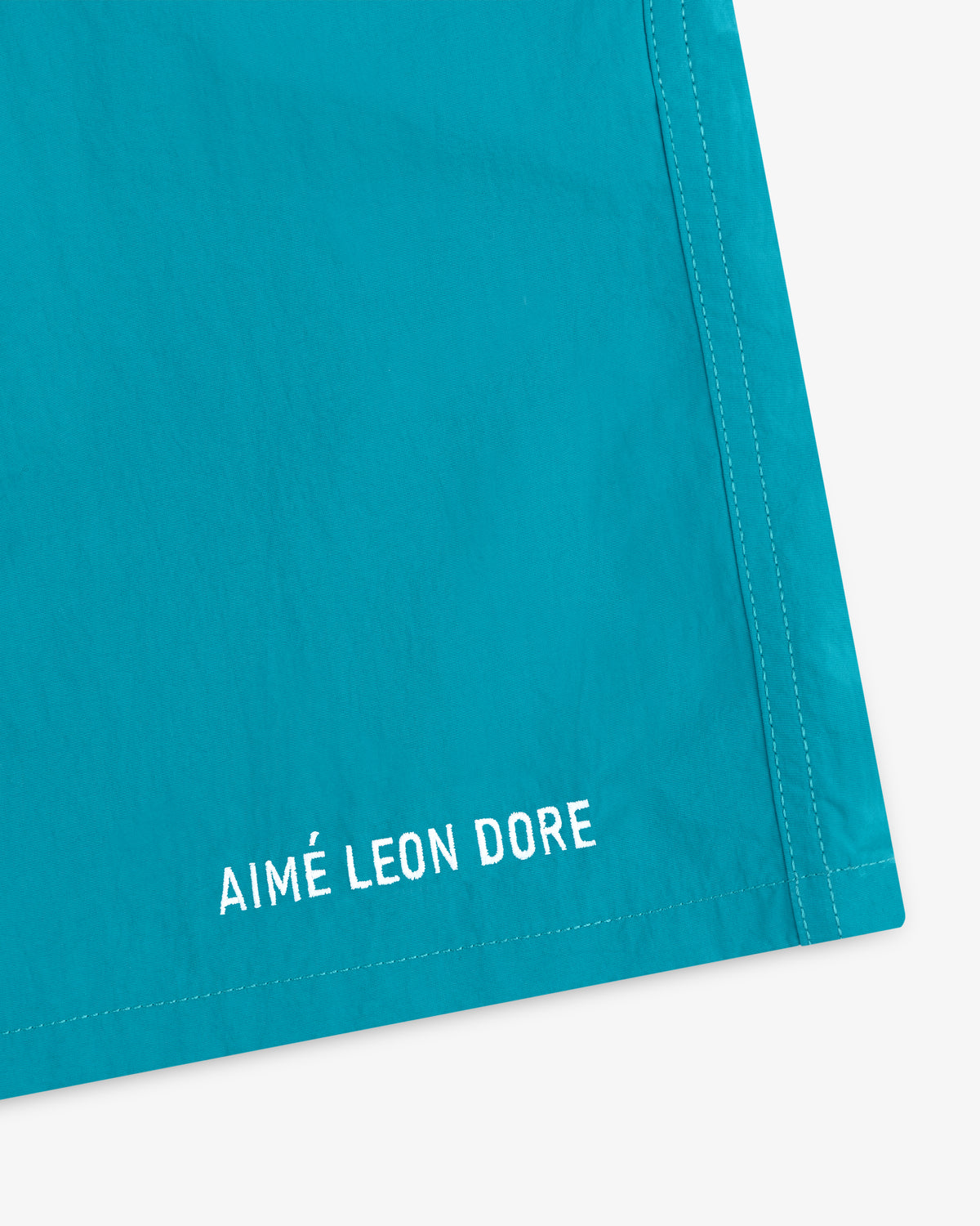 Close-up of turquoise Logo Swim Short with white AIMÉ LEON DORE text embroidered near the hem.