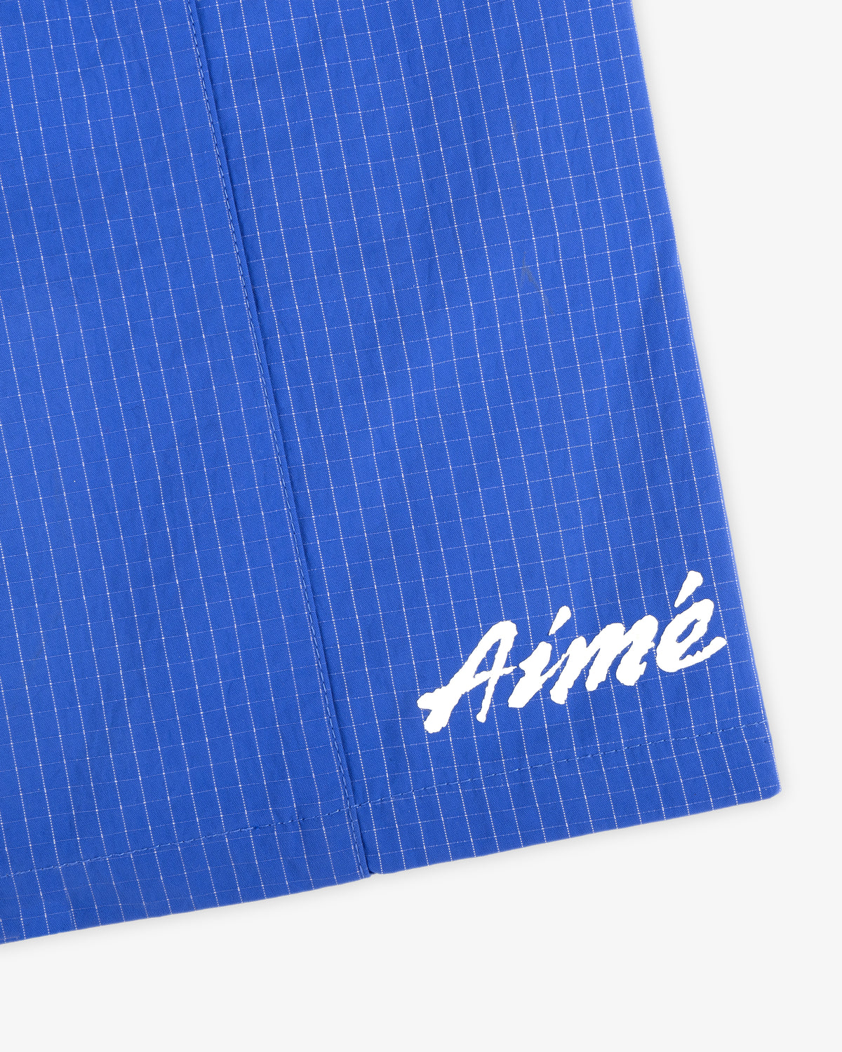 Close-up of blue Ripstop Nylon Wave Short with white grid lines and "Aimé" in white script at the bottom right.