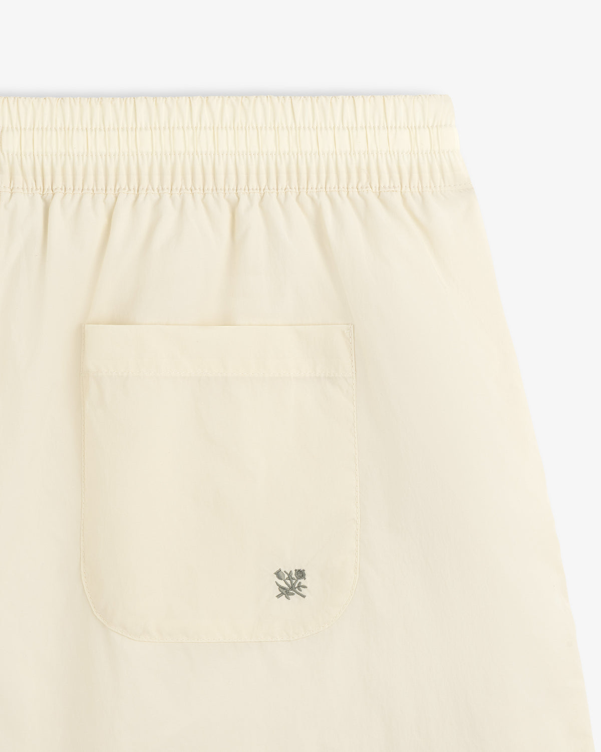 Close-up of Long Nylon Track Short in light beige with an elastic waistband, back pocket, and embroidered logo by the pocket.