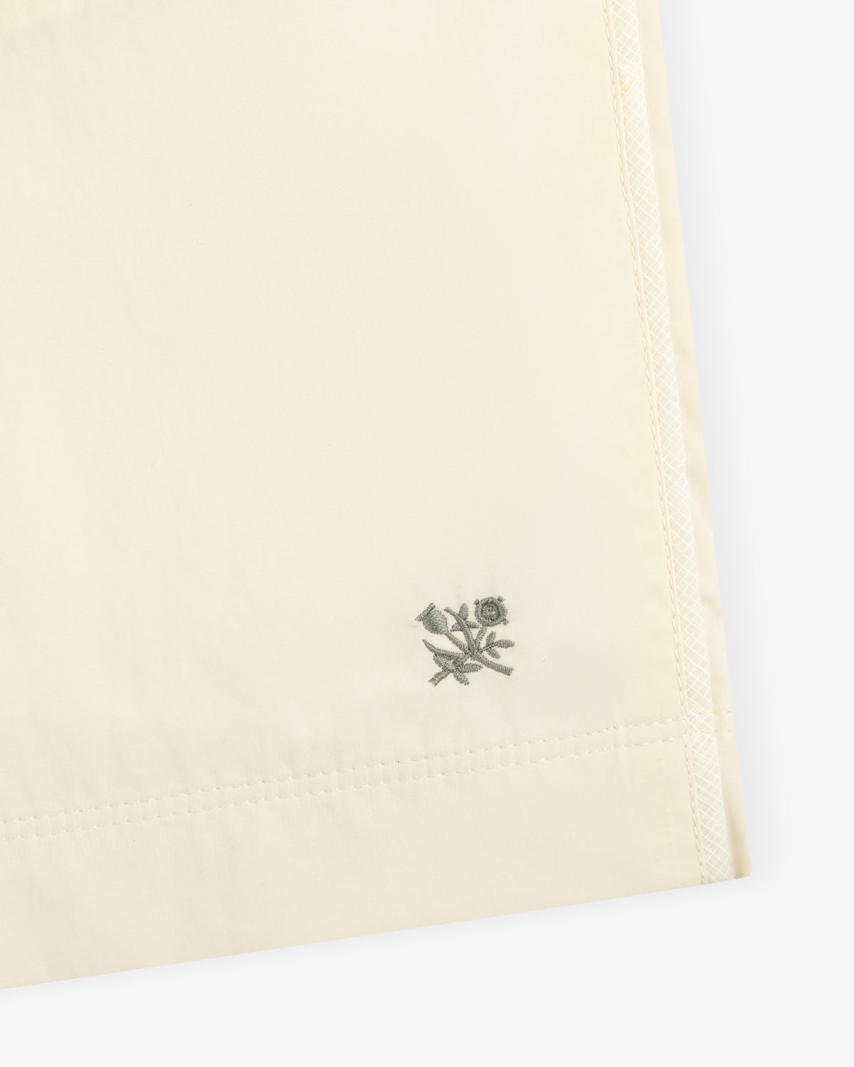 Close-up of Long Nylon Track Short in cream with a small gray embroidered flower and leaf at the corner.