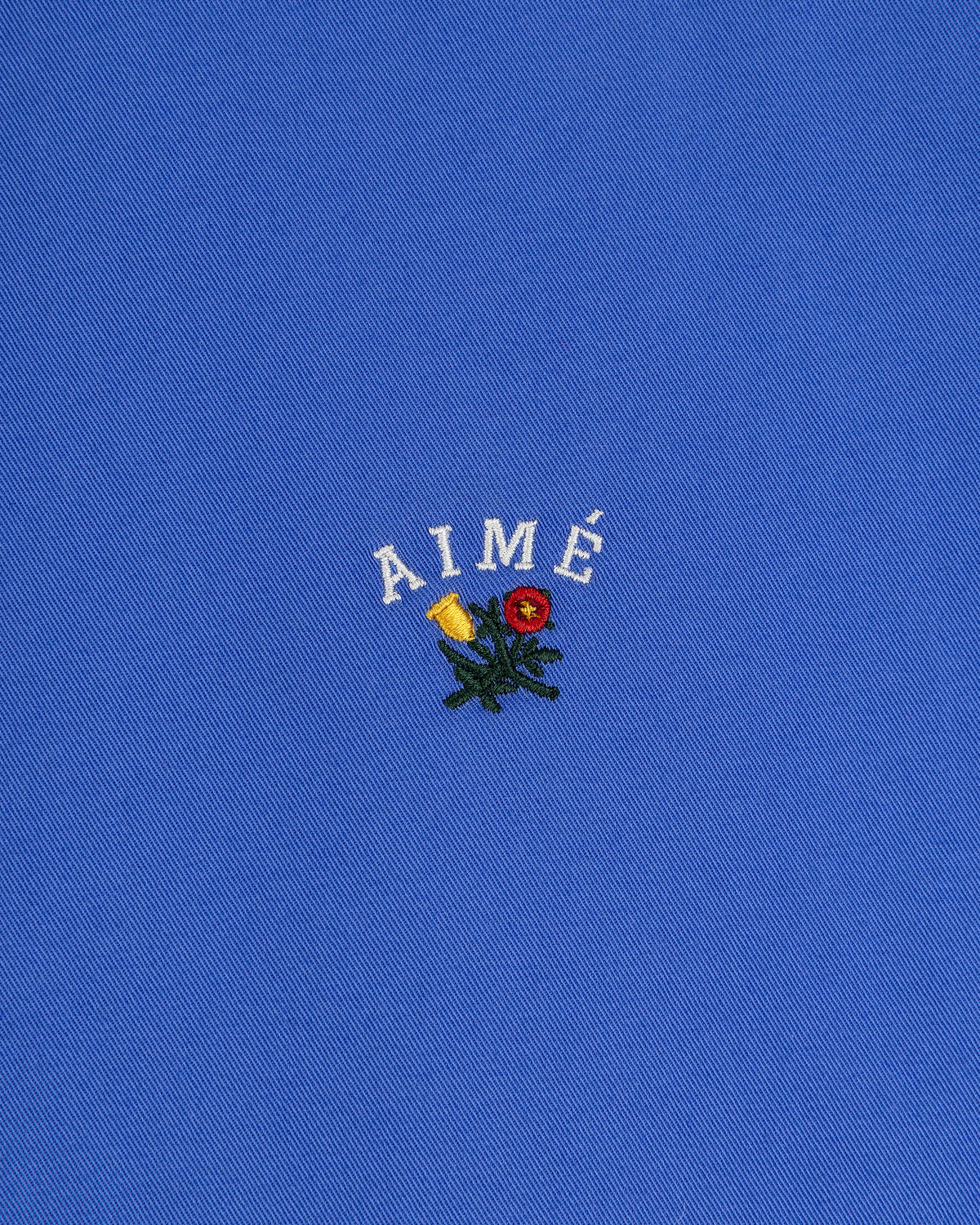 Leon Overshirt: blue fabric with AIMÉ embroidered in white above small yellow and red flowers with green leaves.