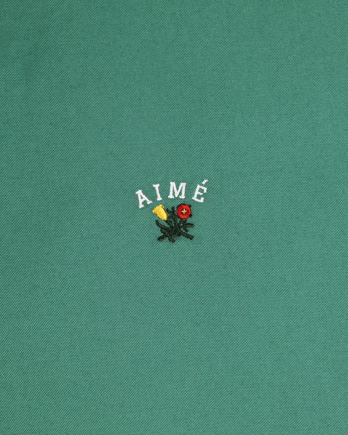 The Leon Overshirt features green fabric with AIMÉ embroidered above yellow and red flowers with green leaves.