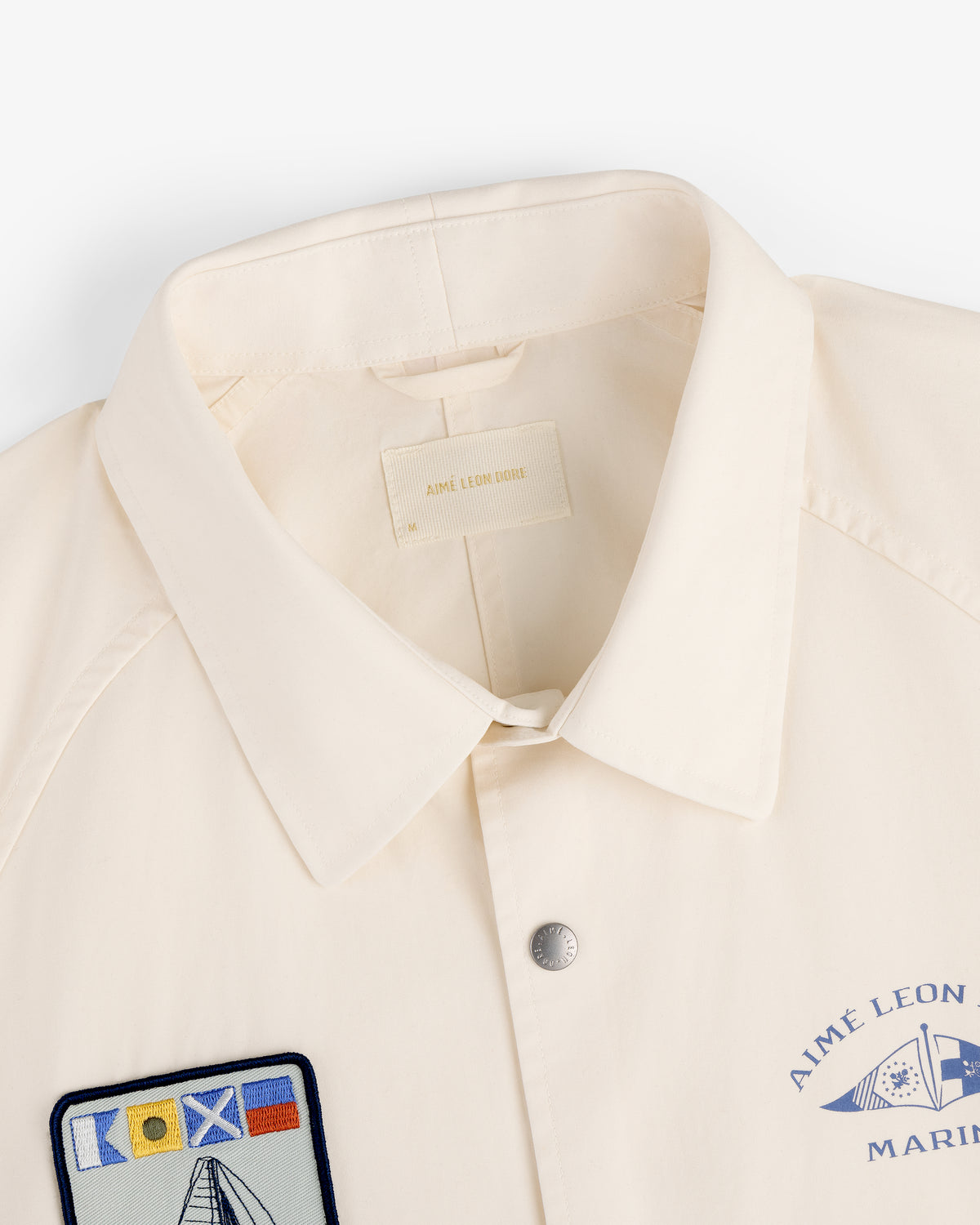 The Marina Jacket is a cream collared jacket with chest patches, featuring a nautical flag and AIME LEON DORE text.