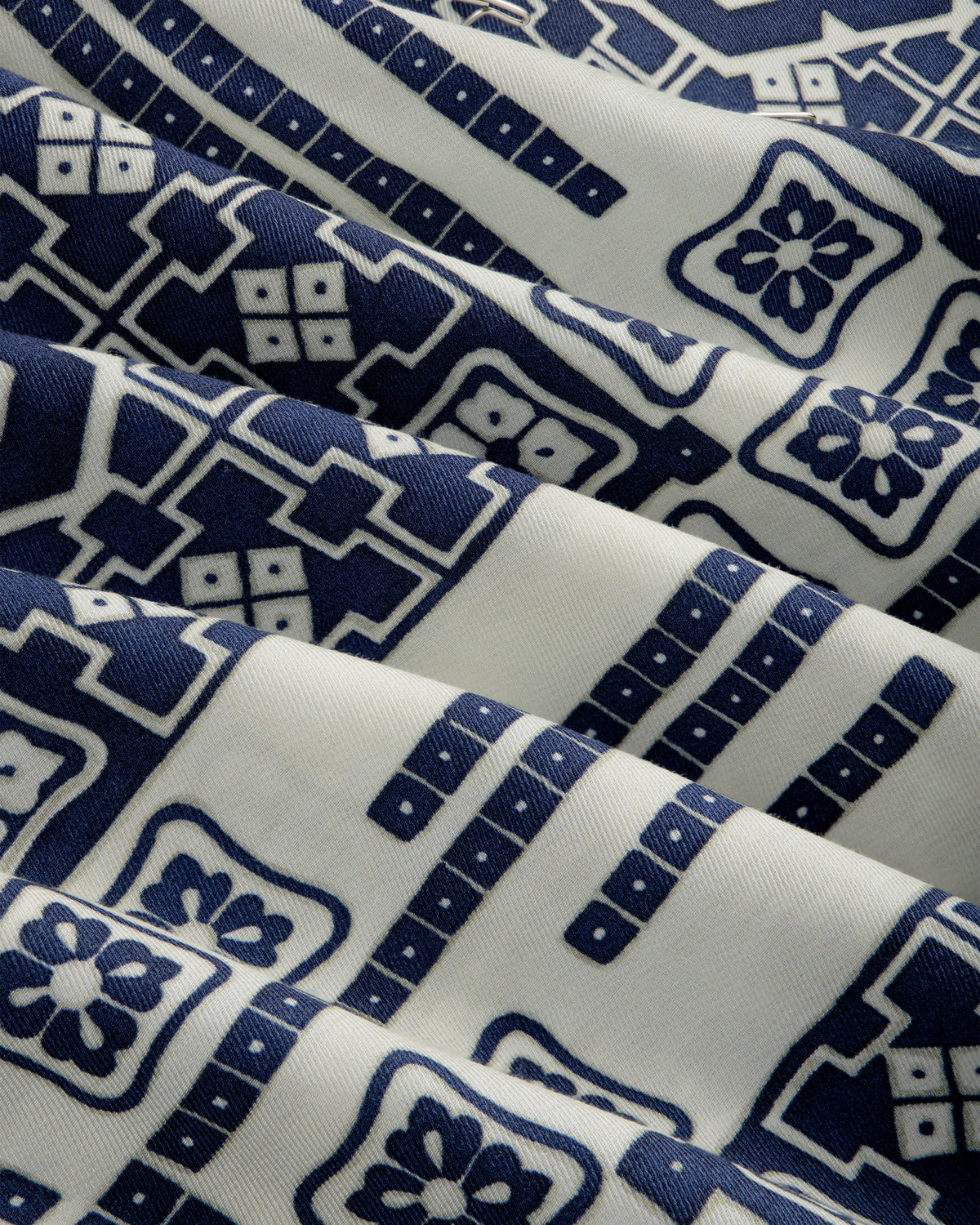 The Mosaic Button Down Shirt features white fabric with blue geometric and floral patterns, shown with gentle folds.