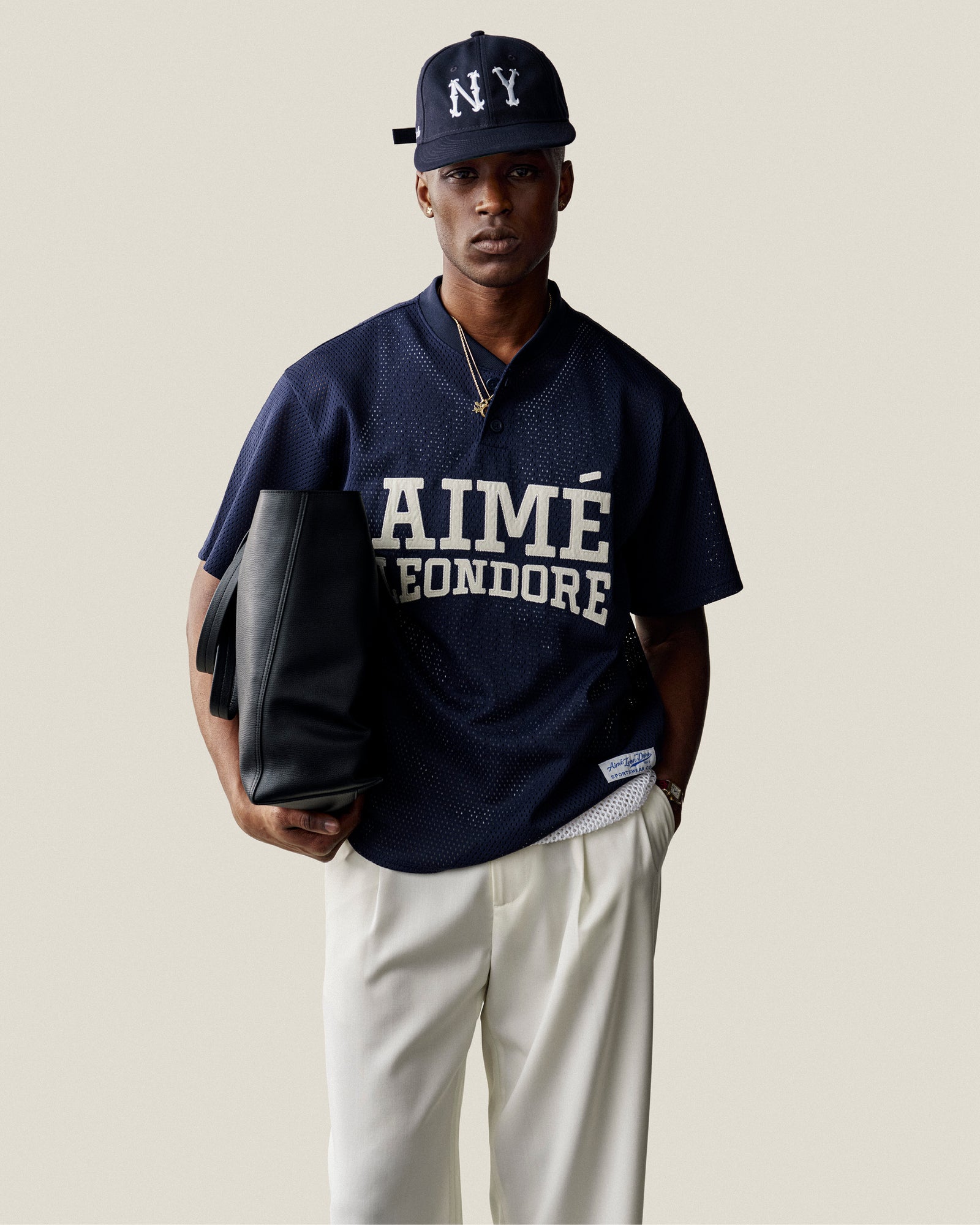 Man in a navy Mesh Batting Practice Jersey, NY cap, white pants, with a black tote bag against a light background.