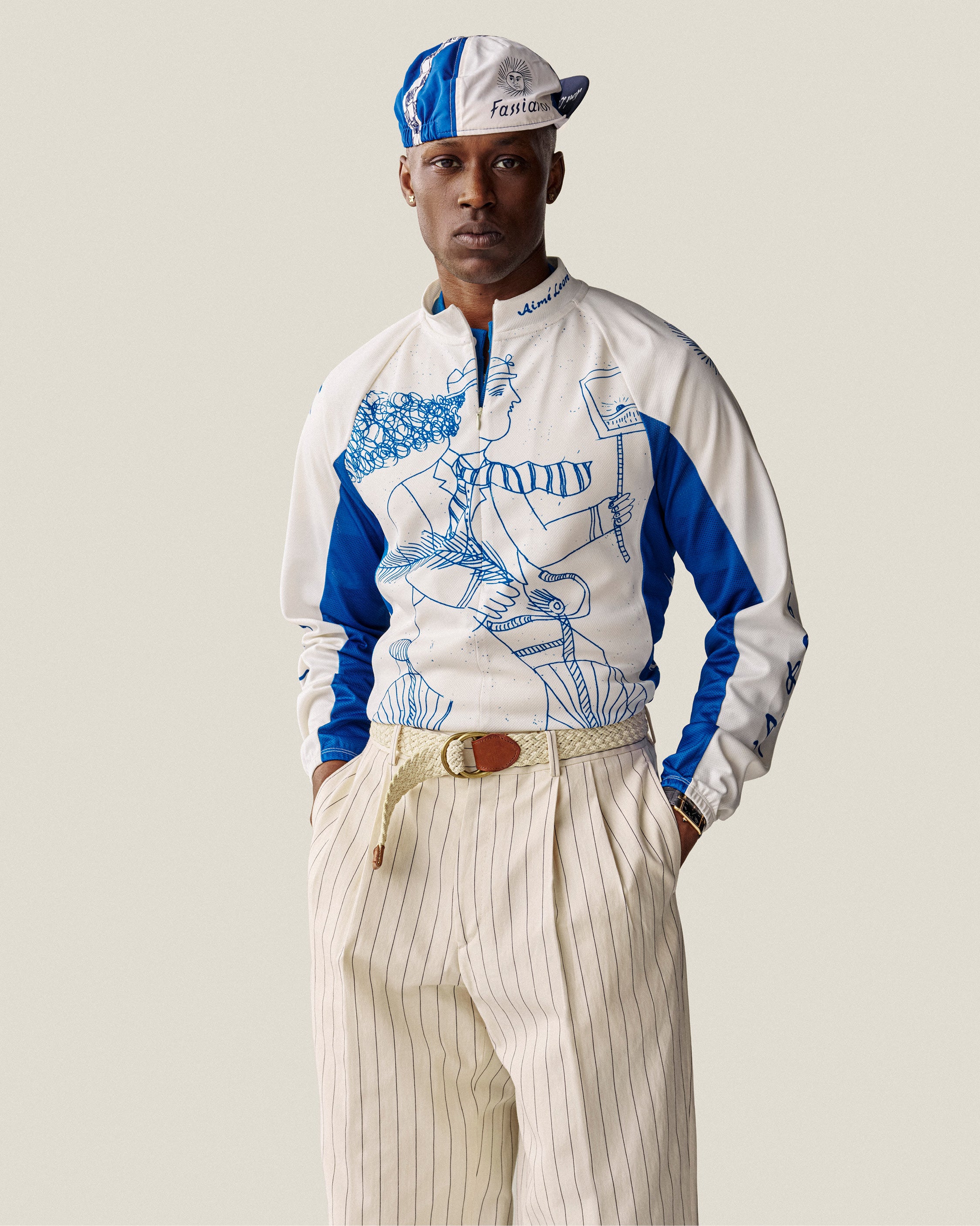 Man wearing a Fassianos Cycling Cap, graphic zip-up top, and beige pinstripe pants with hands in pockets.