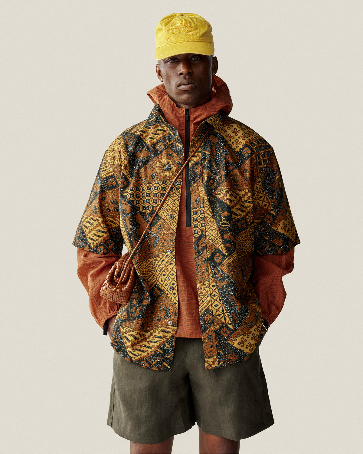 A person in a patterned jacket, orange shirt, yellow cap, olive shorts, and a Woven Leather Shoulder Bag stands against a plain background.