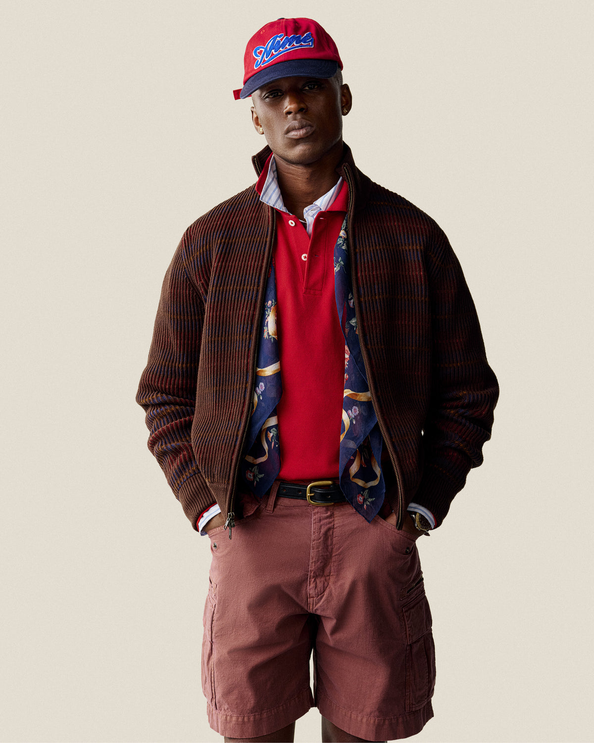 A man in a red cap and Multi Color Knit Track Sweater stands against a plain background.