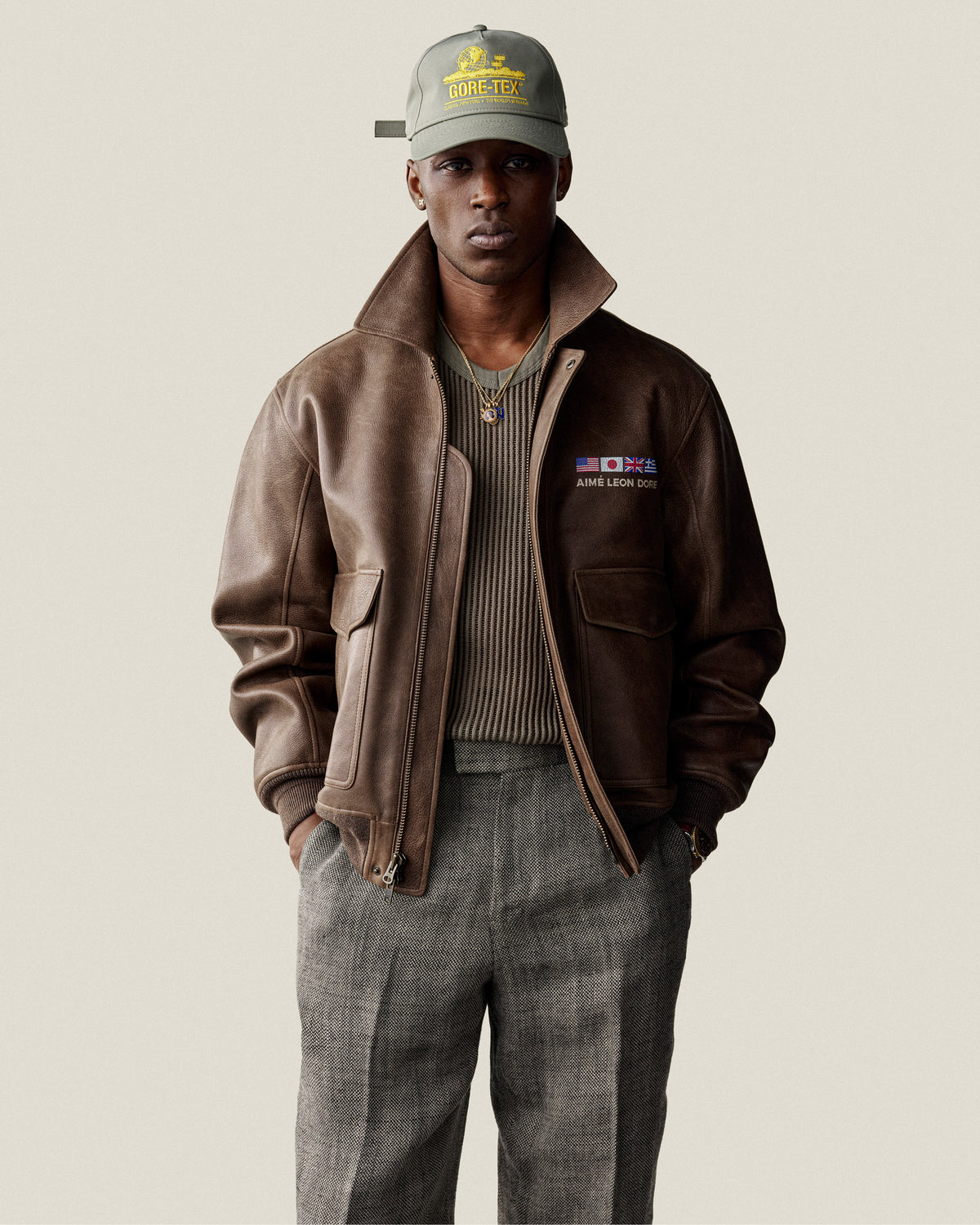 A man in a Mesh V-Neck Tee, gray pants, and a green GORE-TEX cap stands against a plain background.
