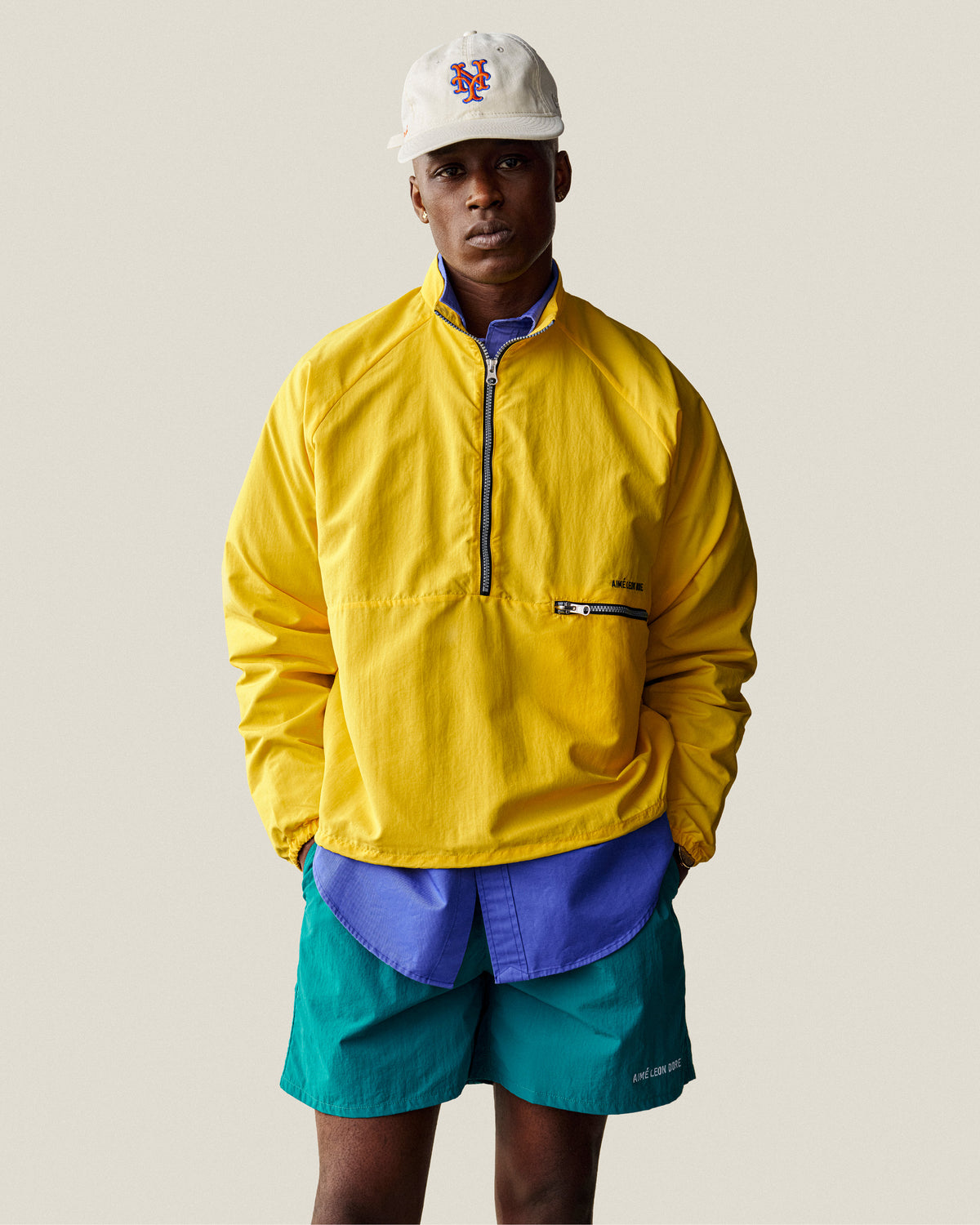 Man in a yellow jacket, teal shorts, blue shirt, and ALD / New Era Mets Retro Fit Hat stands against a plain background.