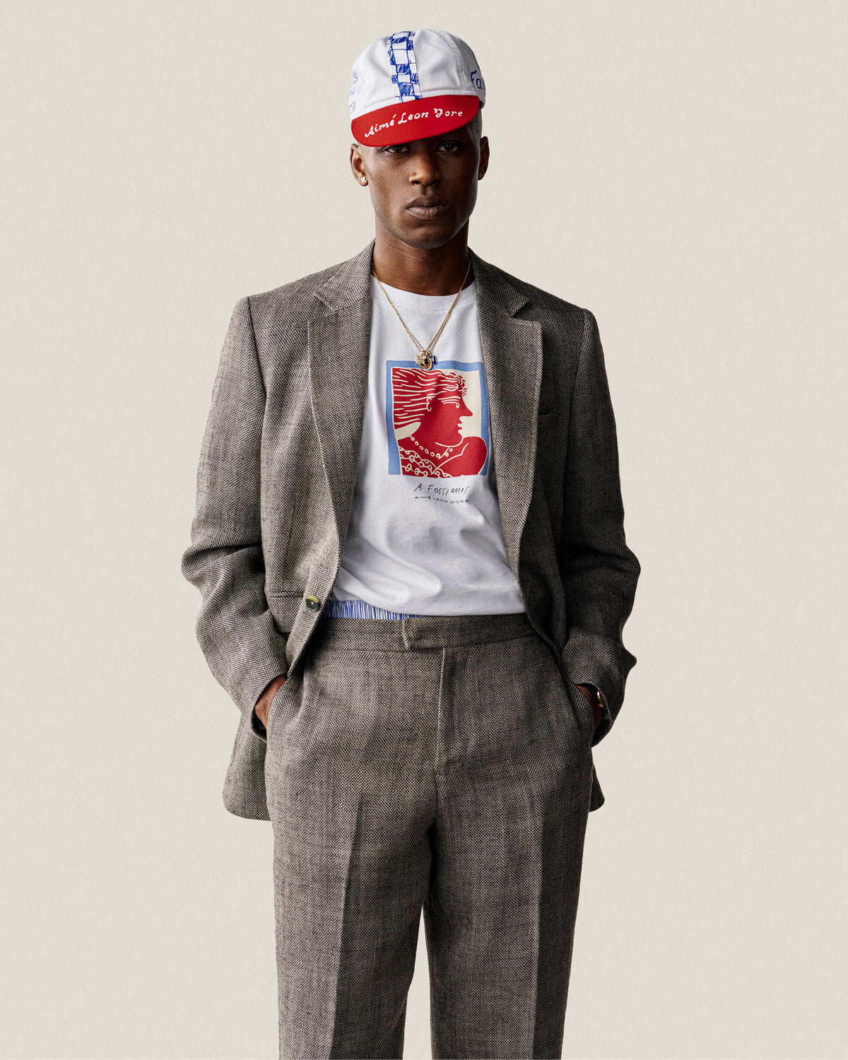 Man wearing Yarn Dyed Linen Suit Trouser, gray suit, white graphic tee, and red-white cap, hands in pockets.