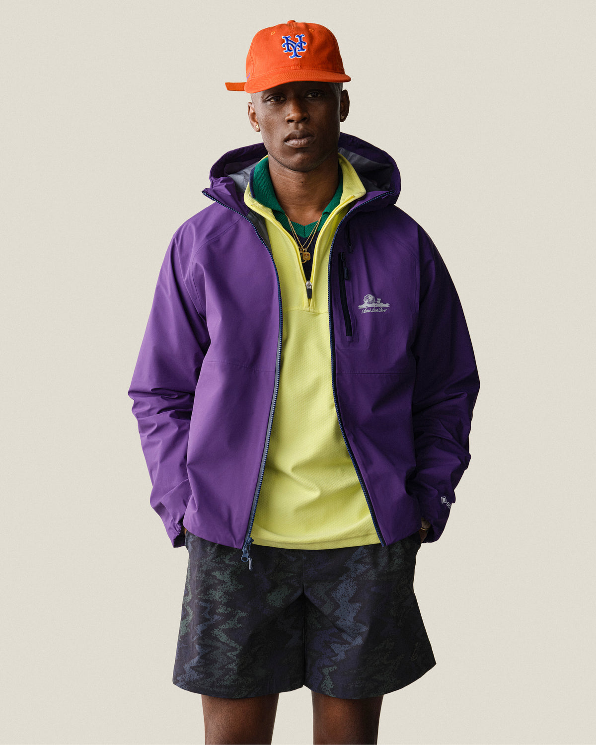 Man wears a purple jacket, yellow shirt, ALD/New Era Mets Retro Fit Hat (orange), and black shorts against a plain background.