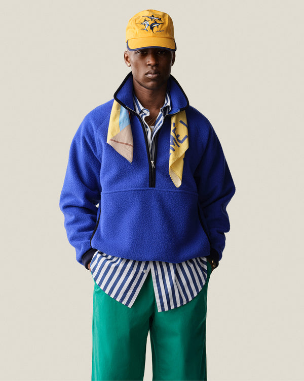 Man in Casentino Crest Half-Zip Pullover, green pants, striped shirt, yellow cap, and yellow scarf against plain background.