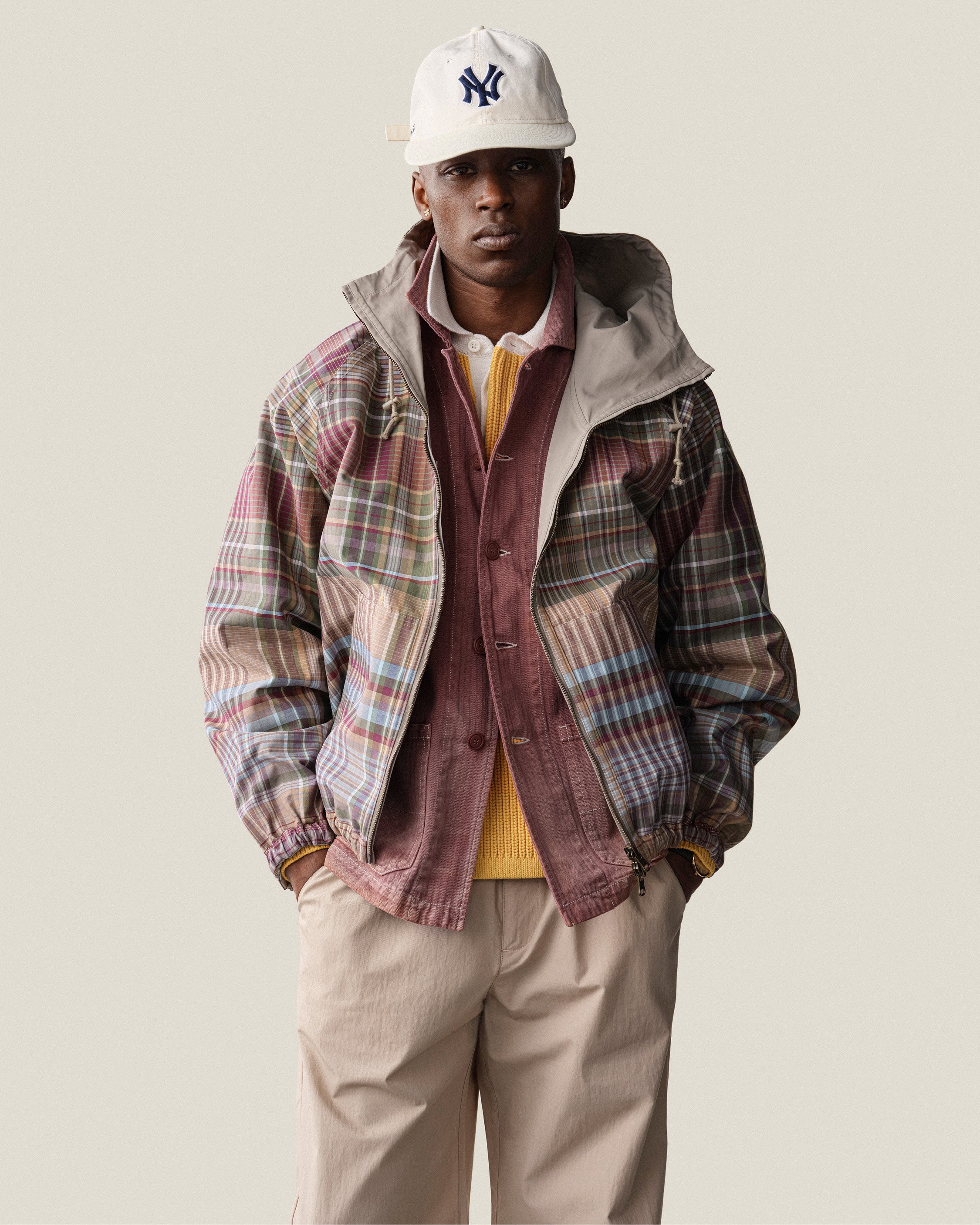 A man wears a plaid jacket, maroon shirt, yellow sweater, beige pants, and an ALD / New Era Yankees Retro Fit Hat.