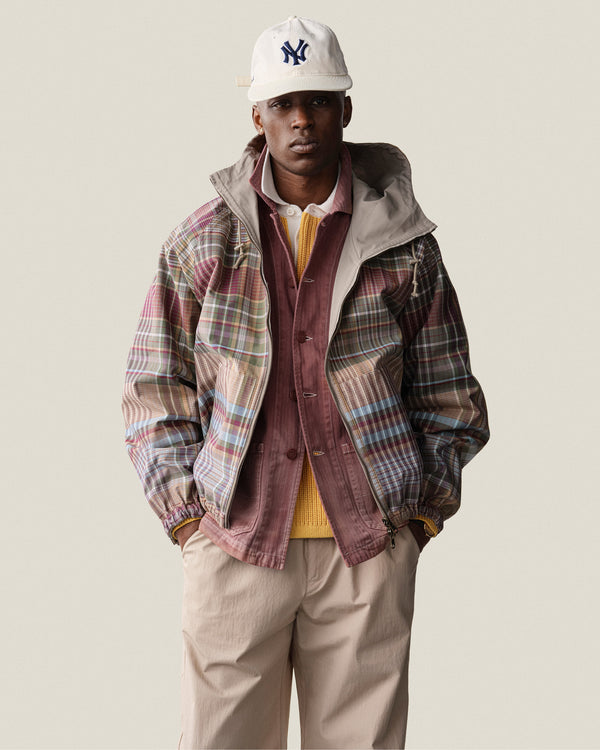 A man wears a plaid jacket, maroon shirt, yellow sweater, beige pants, and an ALD / New Era Yankees Retro Fit Hat.