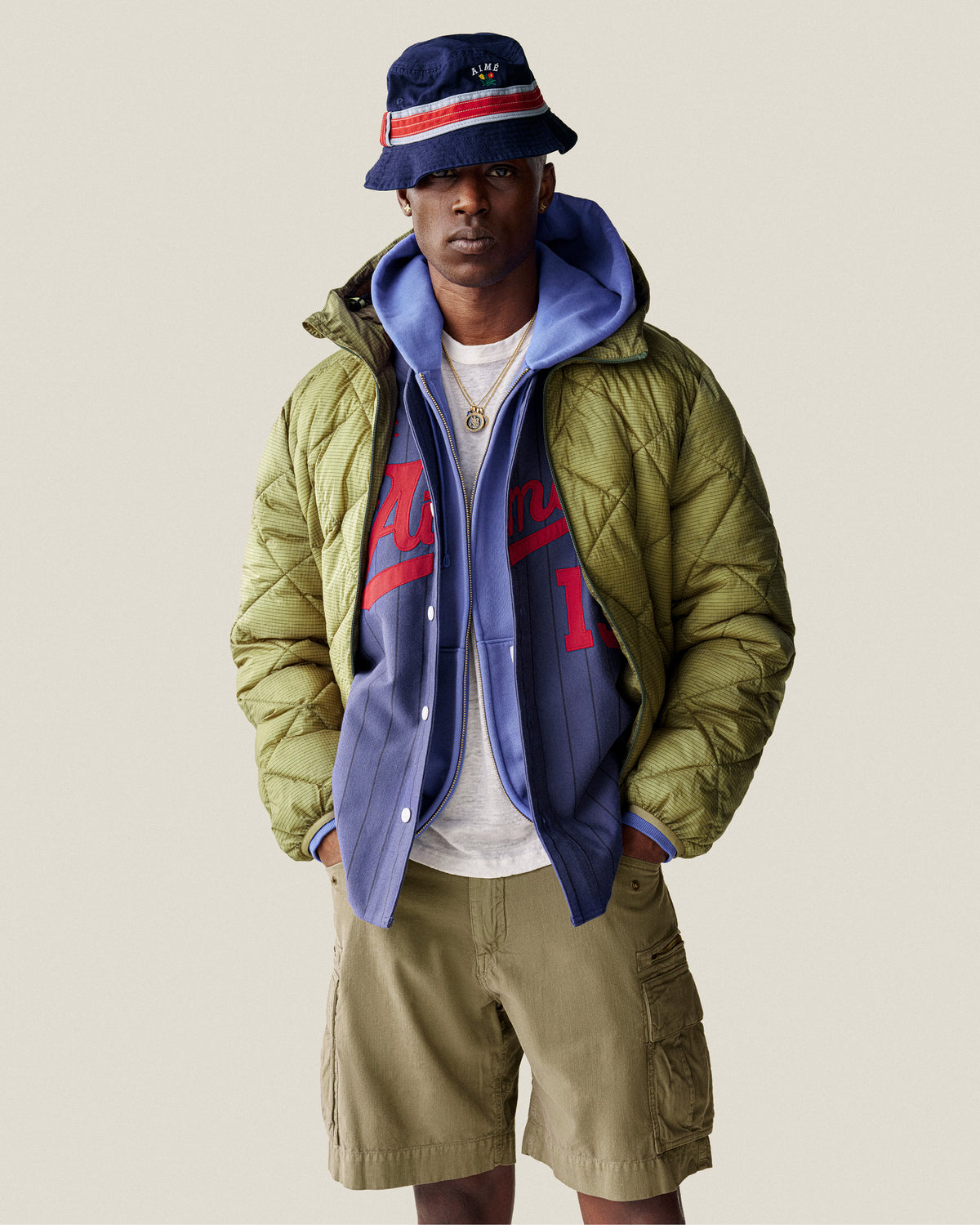 A man wearing a bucket hat, green puffer jacket, Sun Faded Full-Zip Hoodie, sports jersey, and khaki cargo shorts.