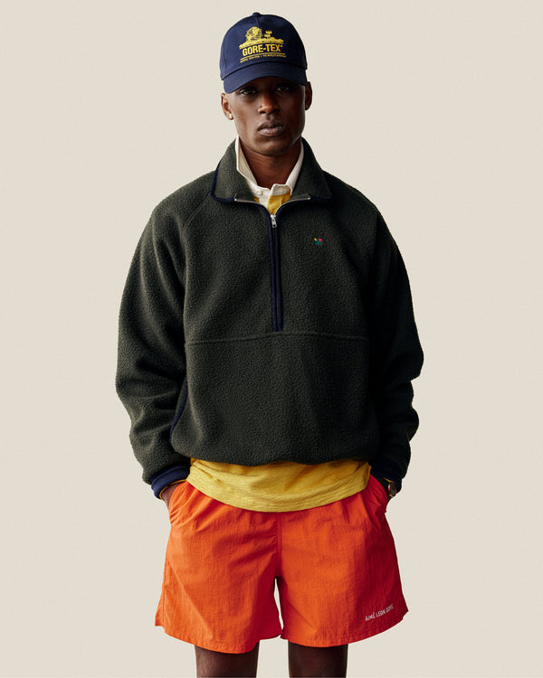 Man wearing bright orange Logo Swim Short, navy cap, dark fleece pullover, and yellow shirt, hands in pockets.