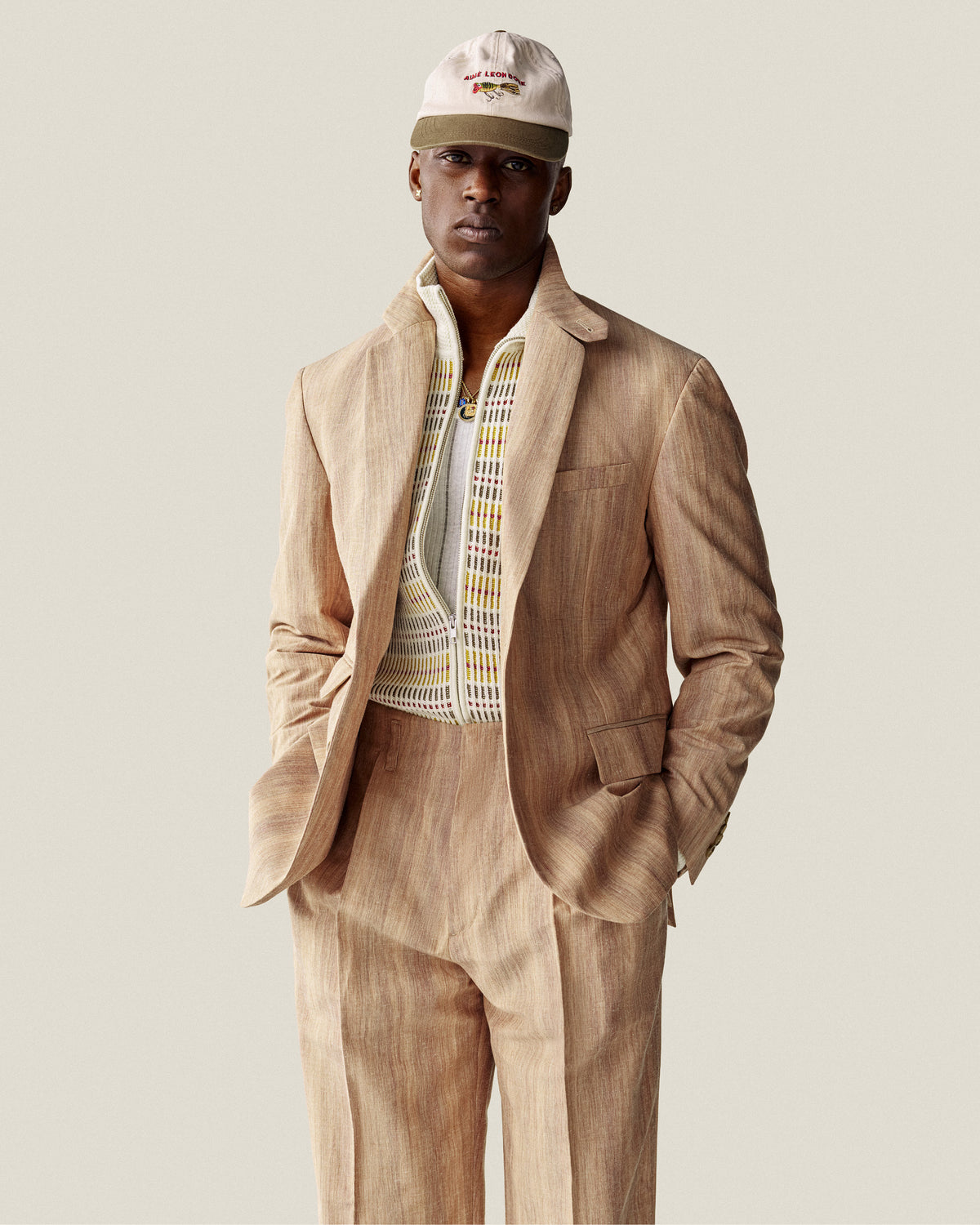 Man in Multi Color Knit Track Sweater and white cap, standing with hands in pockets against a plain background.