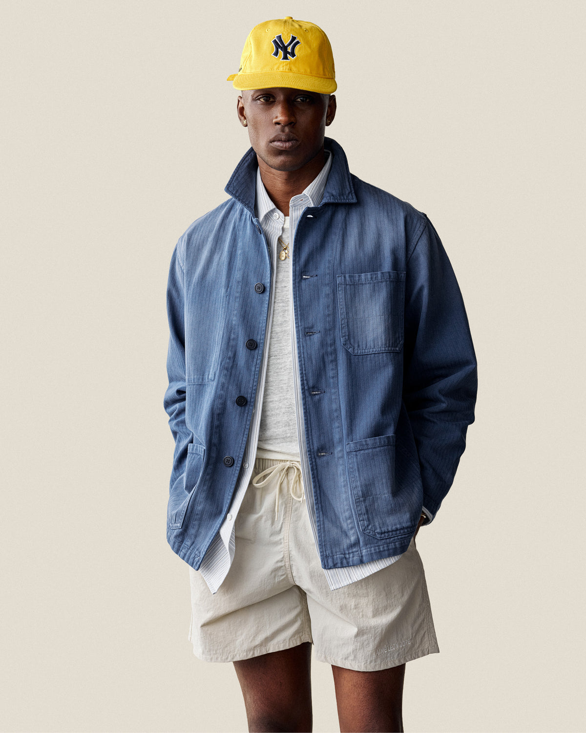 Man wears an ALD / New Era Yankees Retro Fit Hat, blue jacket, gray shirt, and beige shorts against a plain background.