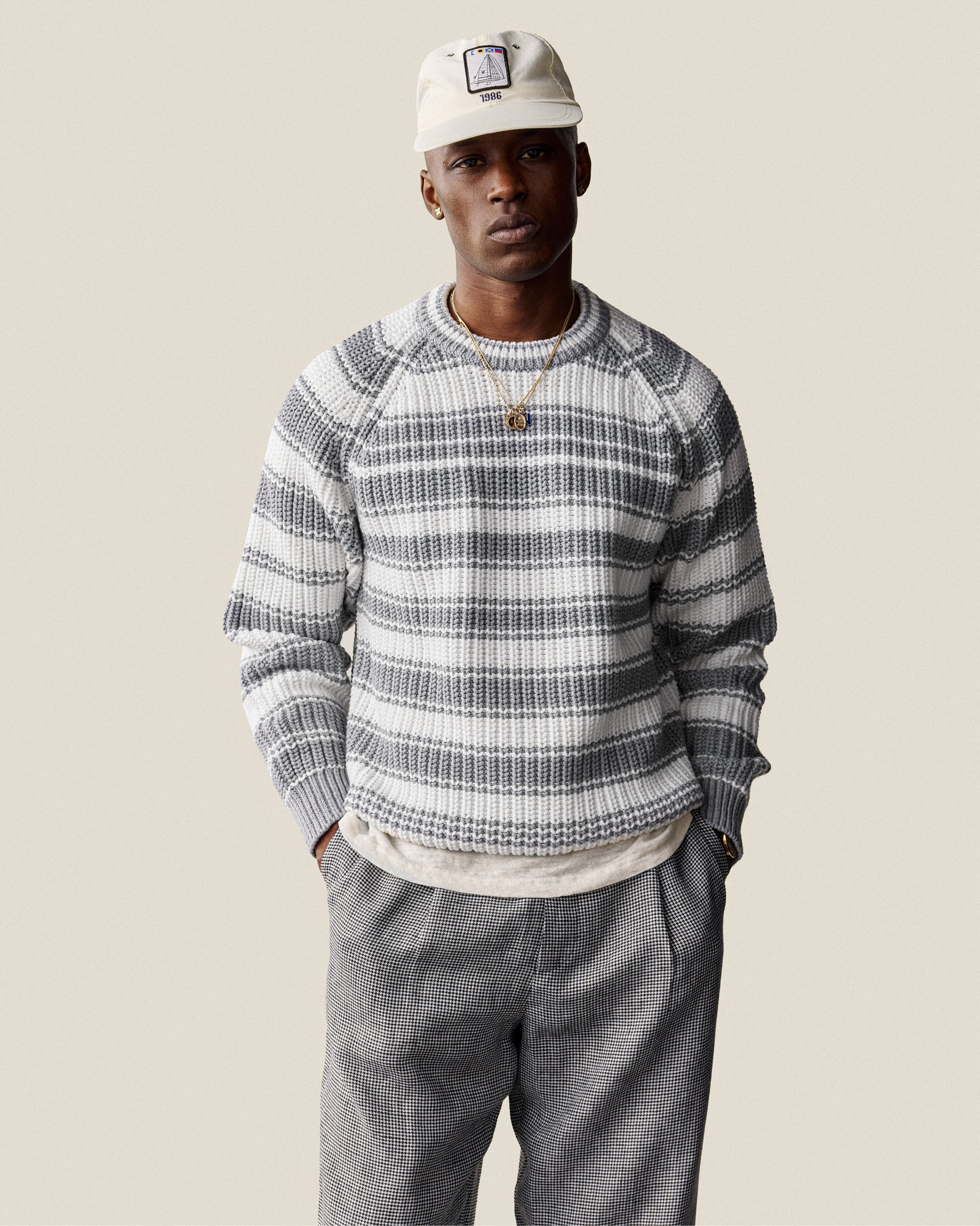 Man in a Striped Crewneck Sweater, gray pants, and beige cap stands with hands in pockets against a plain background.