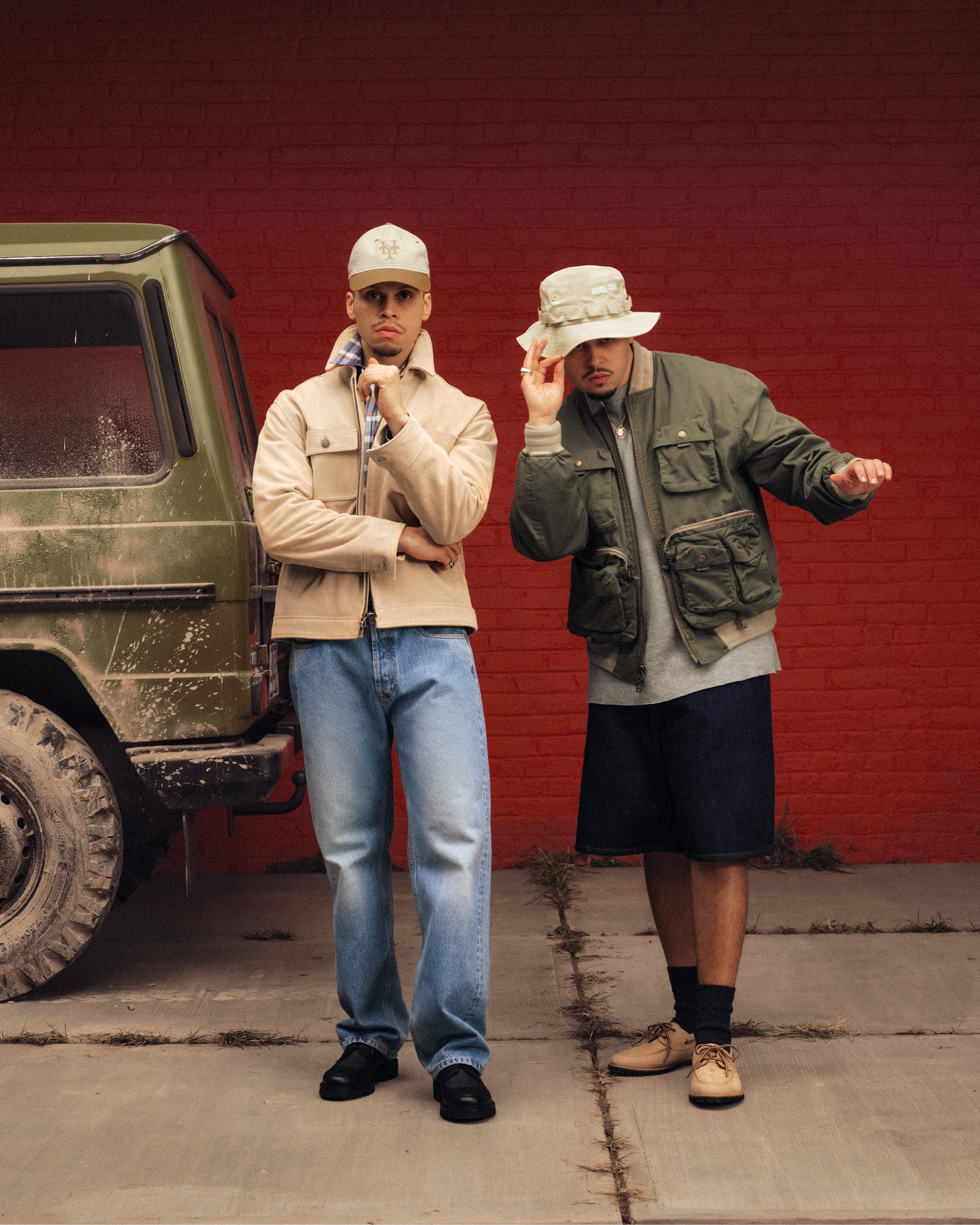 Two men in Leather Ranch Jackets and hats pose by a muddy green SUV, with a red brick wall in the background.