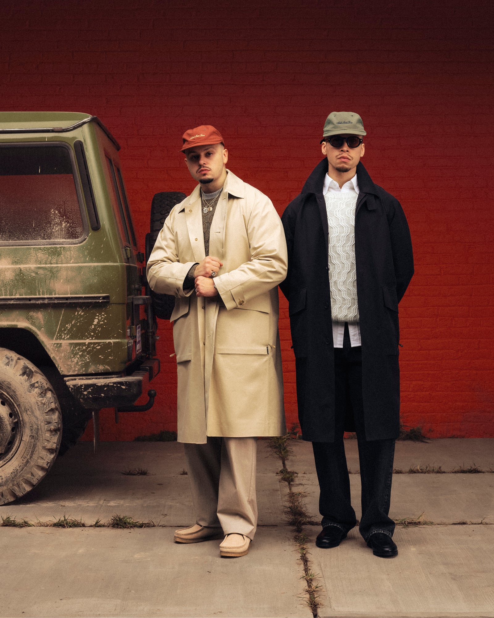 Two men in GORE-TEX Balmacaan Coats and hats stand beside a green SUV by a red brick wall.