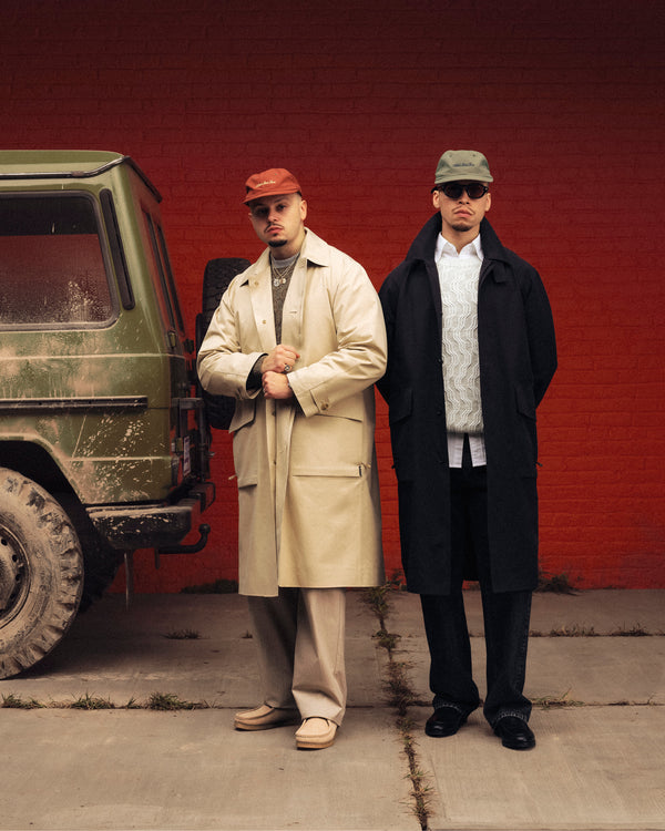 Two men in Linen Blend Cable Knit Sweaters stand by a muddy green SUV against a red brick wall.