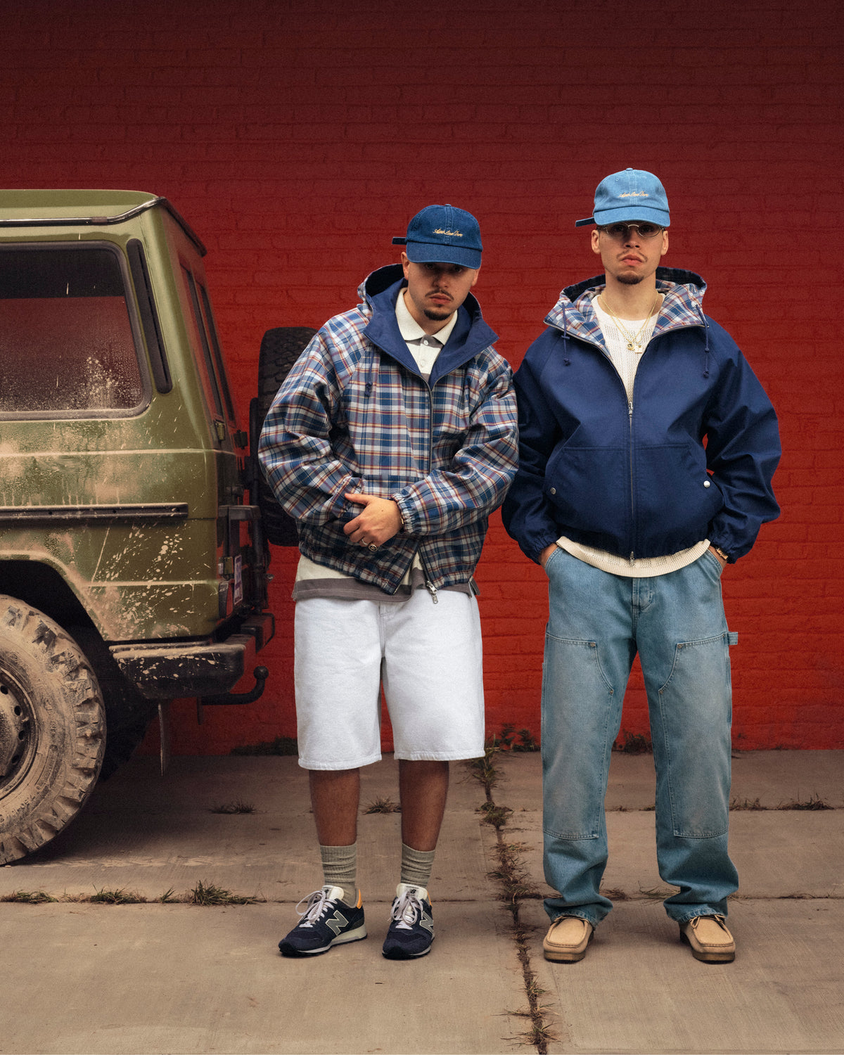 Two men pose by a green SUV and red wall, both wearing blue caps, jackets, and Racking Stitch Crewneck Sweaters.