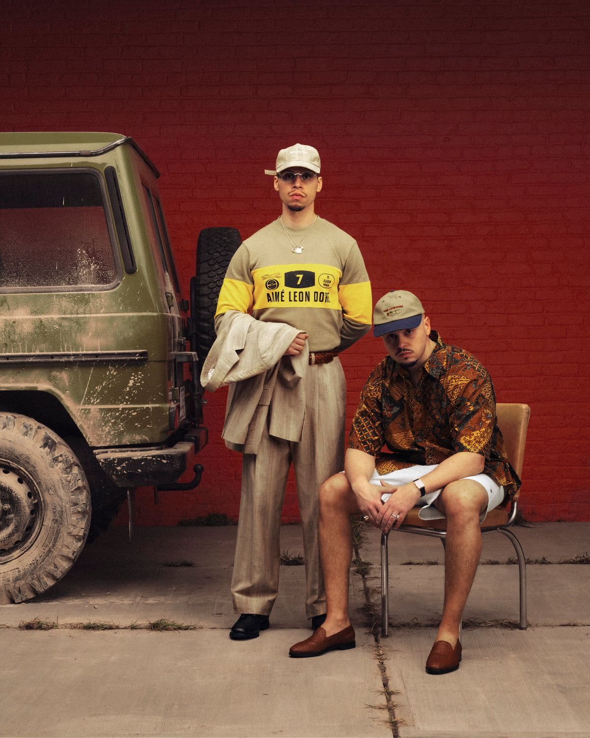 Two men in Long-Sleeve Rally Tees pose by a muddy jeep and red wall; one holds a jacket, the other sits on a chair.