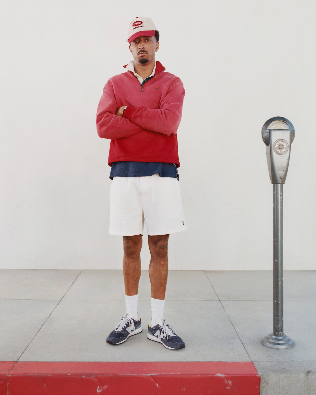 A man in a Sun Faded Crest Quarter Zip, white shorts, and sneakers stands by a parking meter with arms crossed.