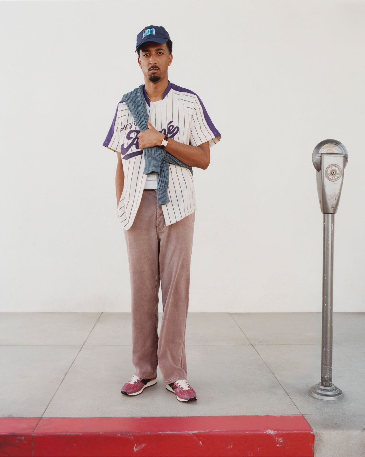 A man in an Aimé Baseball Jersey and cap stands by a parking meter, holding a sweater over his shoulder.