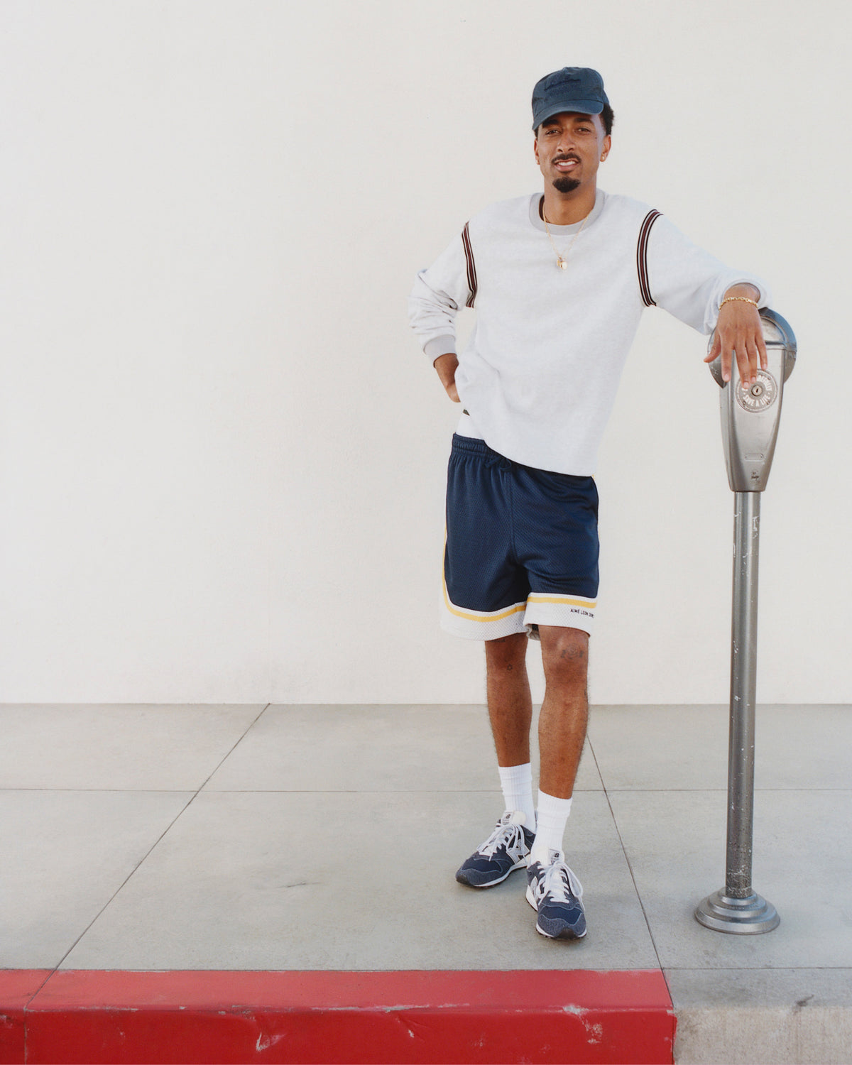 A man in a Long-Sleeve Slub Football Tee, cap, and shorts stands by a parking meter on a sidewalk.